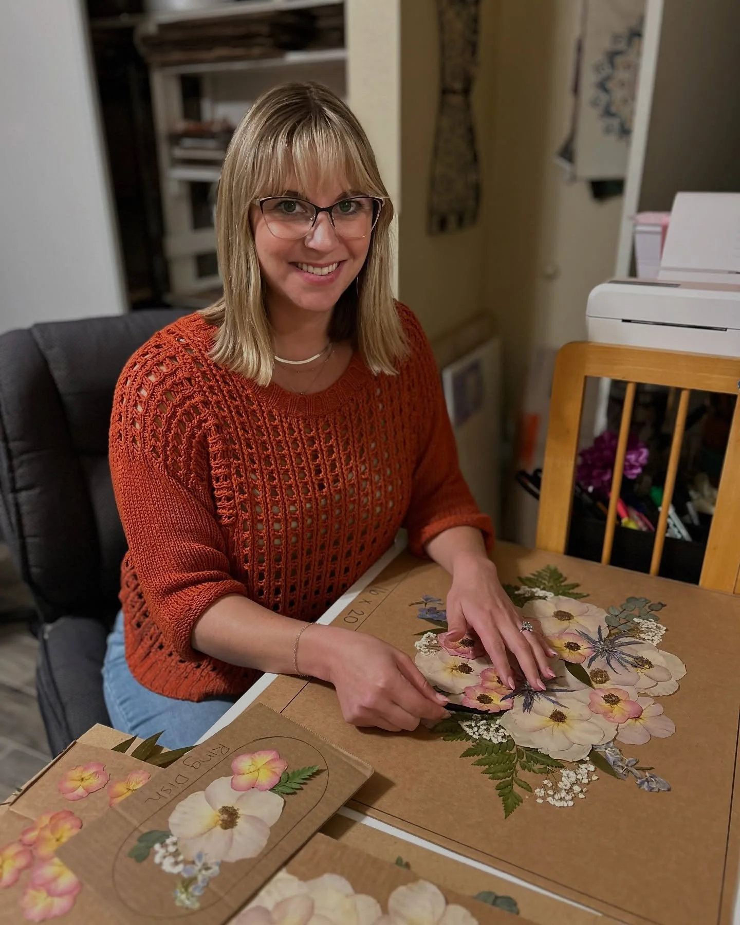 A woman with blonde hair, glasses, and a warm smile sitting at a table, working on a floral collage craft with paper flowers.