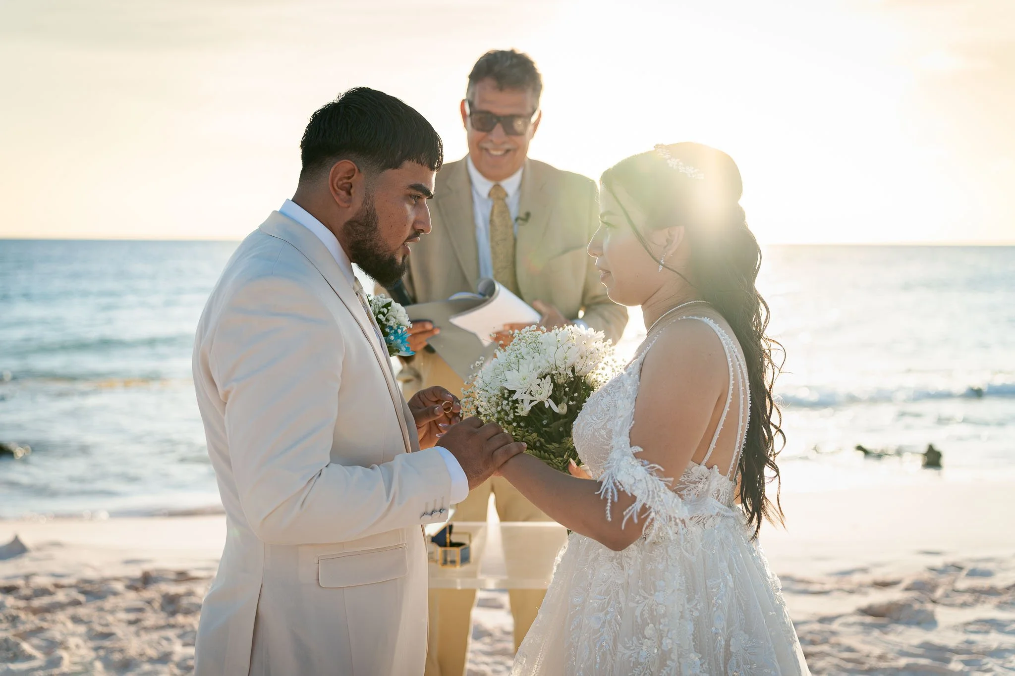 Een paar dat trouwt op het strand, de man legt de ring op de vinger van de vrouw terwijl ze een boekje vasthoudt, de officiant staat lachend op de achtergrond bij zonsondergang.