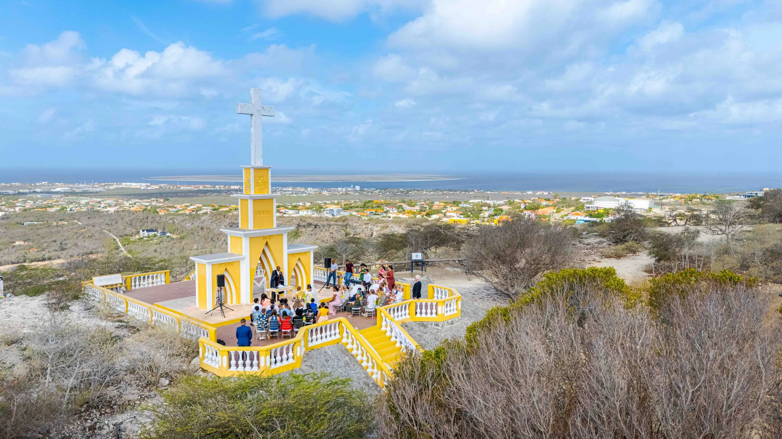 Een groep mensen die een ceremonie bijwoont bij een witgele kapel met een kruis bovenop, gelegen op een heuvel met uitzicht over een stad en de oceaan onder een deels bewolkte hemel.