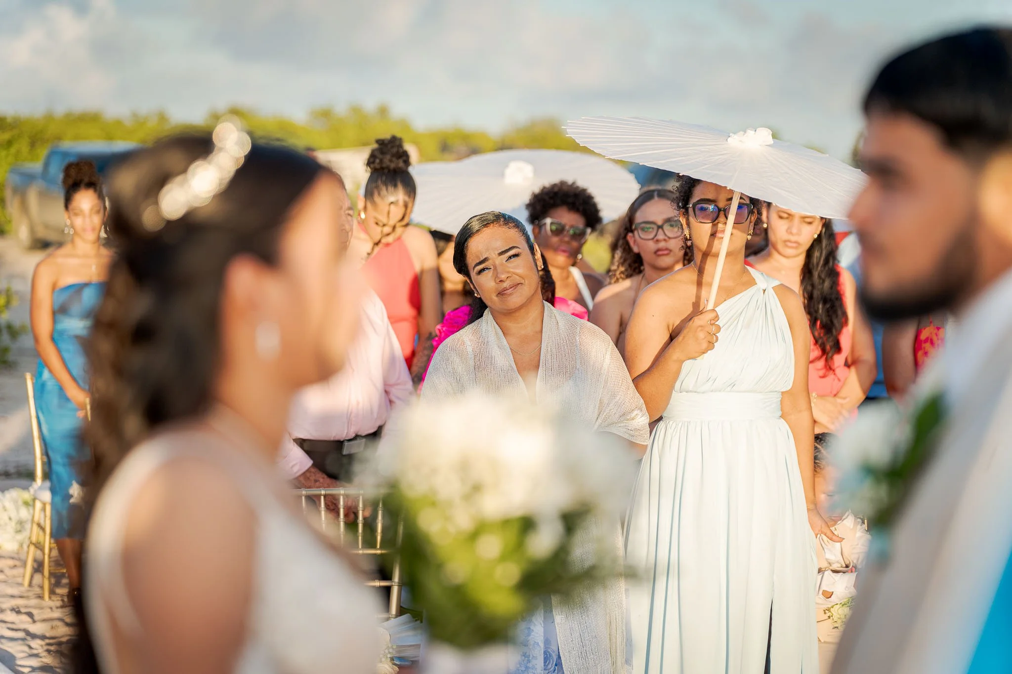 Een groep mensen bij een bruiloft op het strand, met een vrouw die verdrietig lijkt en een woman met een parasol.