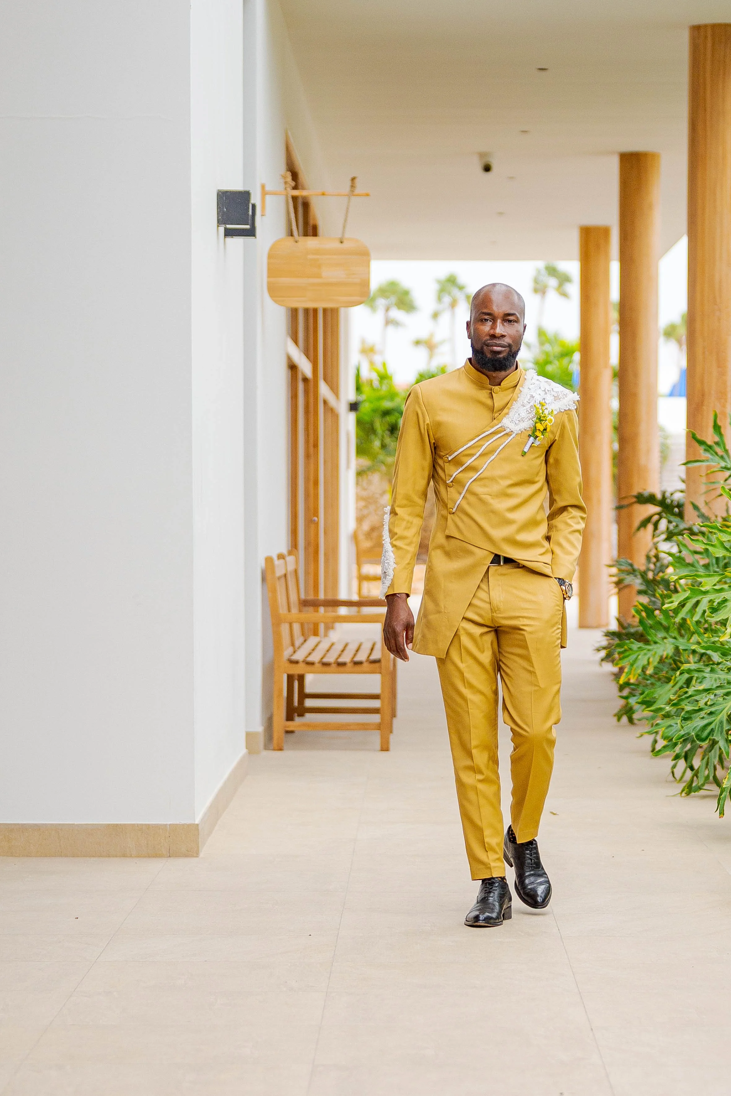 Man in een gouden pak loopt in een corridor met planten en houten meubels.