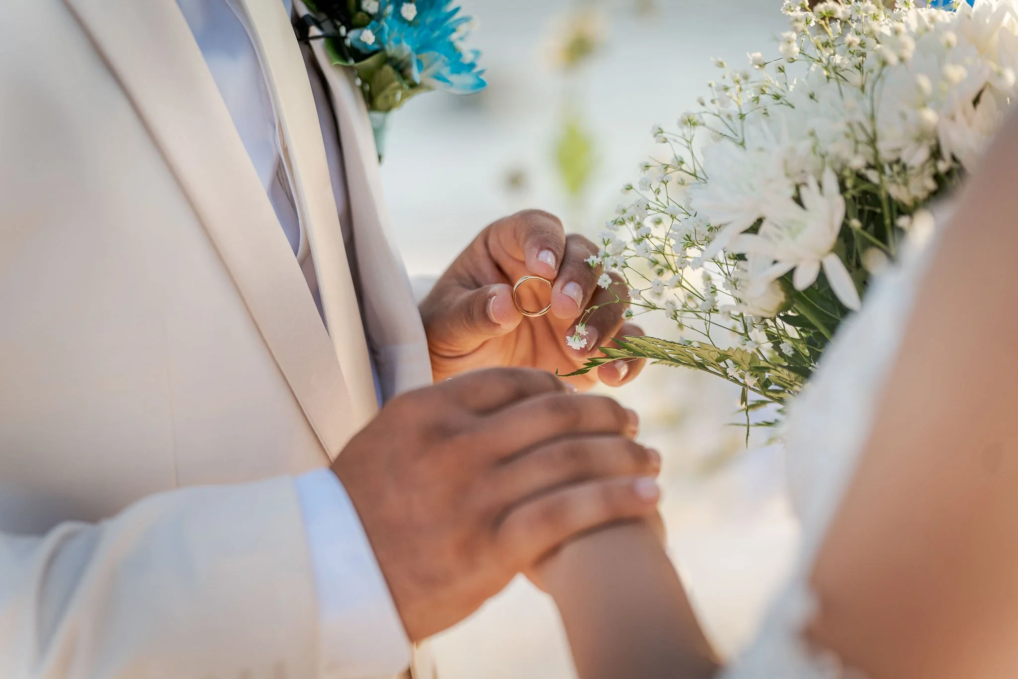 Close-up van een hand die trouwringen vasthoudt, met een persoon die een jurk draagt en een bloemenarrangement vasthoudt op een bruiloft.