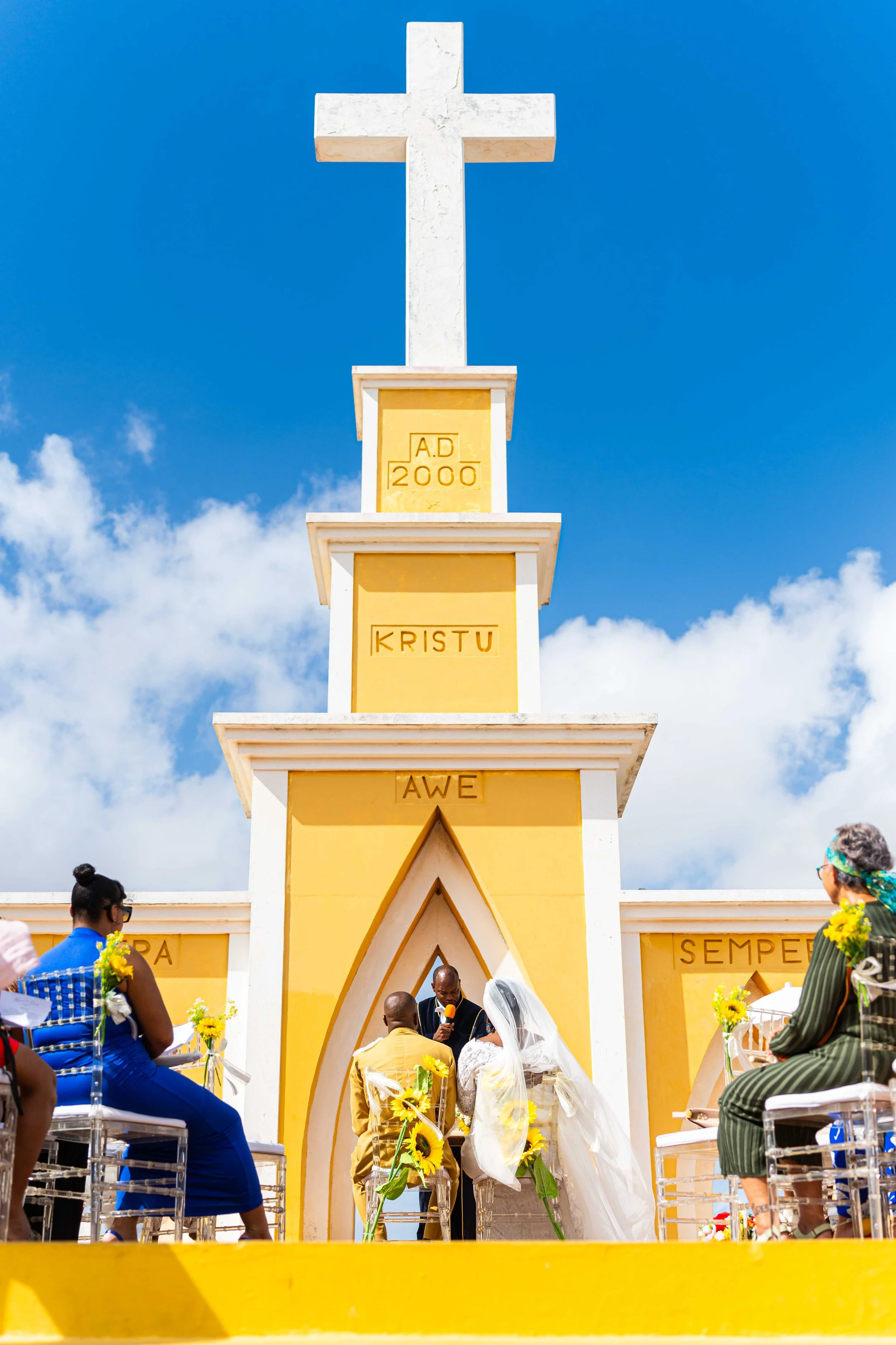 Een huwelijksceremonie voor een kerkgebouw met een grote gele en witte toren en een kruis bovenaan. Mensen zitten op stoelen terwijl een paar voor de dom hangt, omringd door zonnebloemen.