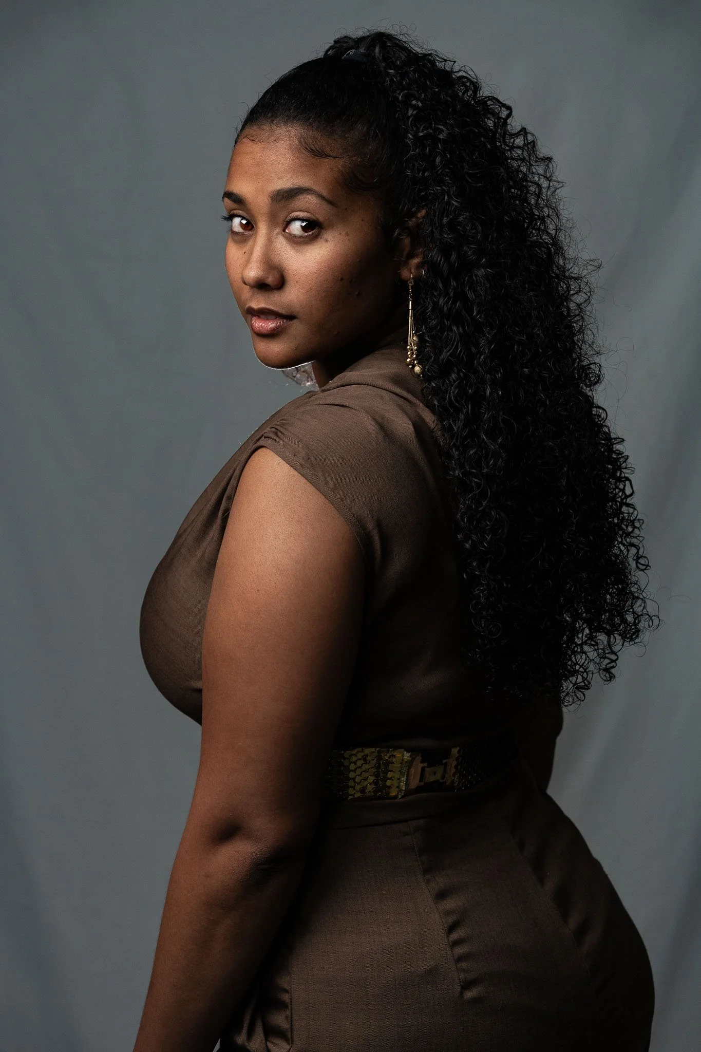 Portret van een jonge vrouw met lang, krullend zwart haar, gekleed in een bruin blouse en rok, in een neutrale grijze achtergrond.