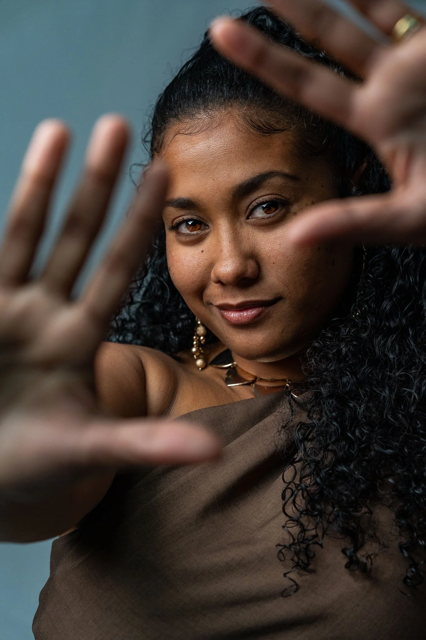 Een jonge vrouw met donkere, gekrulde haar en een zelfverzekerde blik, die naar de camera kijkt en haar hand voor haar gezicht houdt.