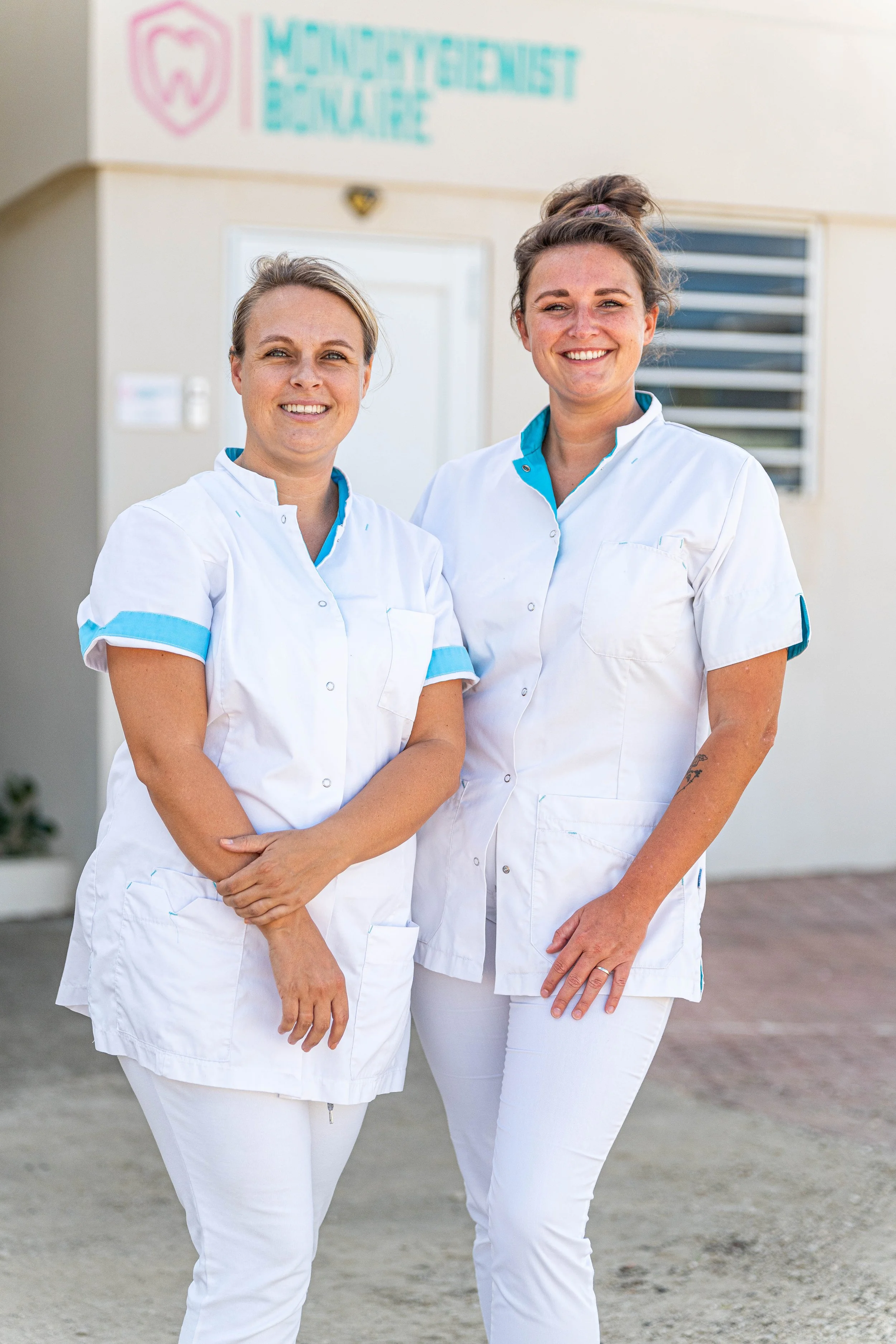 Twee vrouwelijke tandarts of tandheelkundige professionals in witte uniformen staan buiten voor een kliniek of praktijk met een beveiligingscamera en een logo die 'WONENDE INT WANDE' lezen.