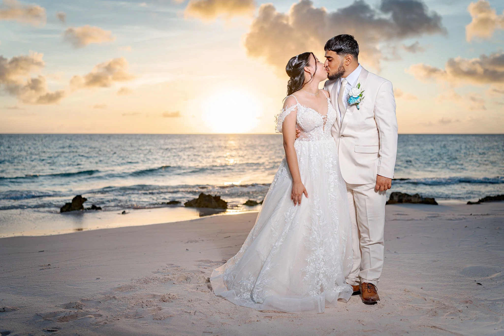 Een getrouwd stel kust op het strand tijdens zonsondergang, de vrouw draagt een witte trouwjurk en de man een wit pak.