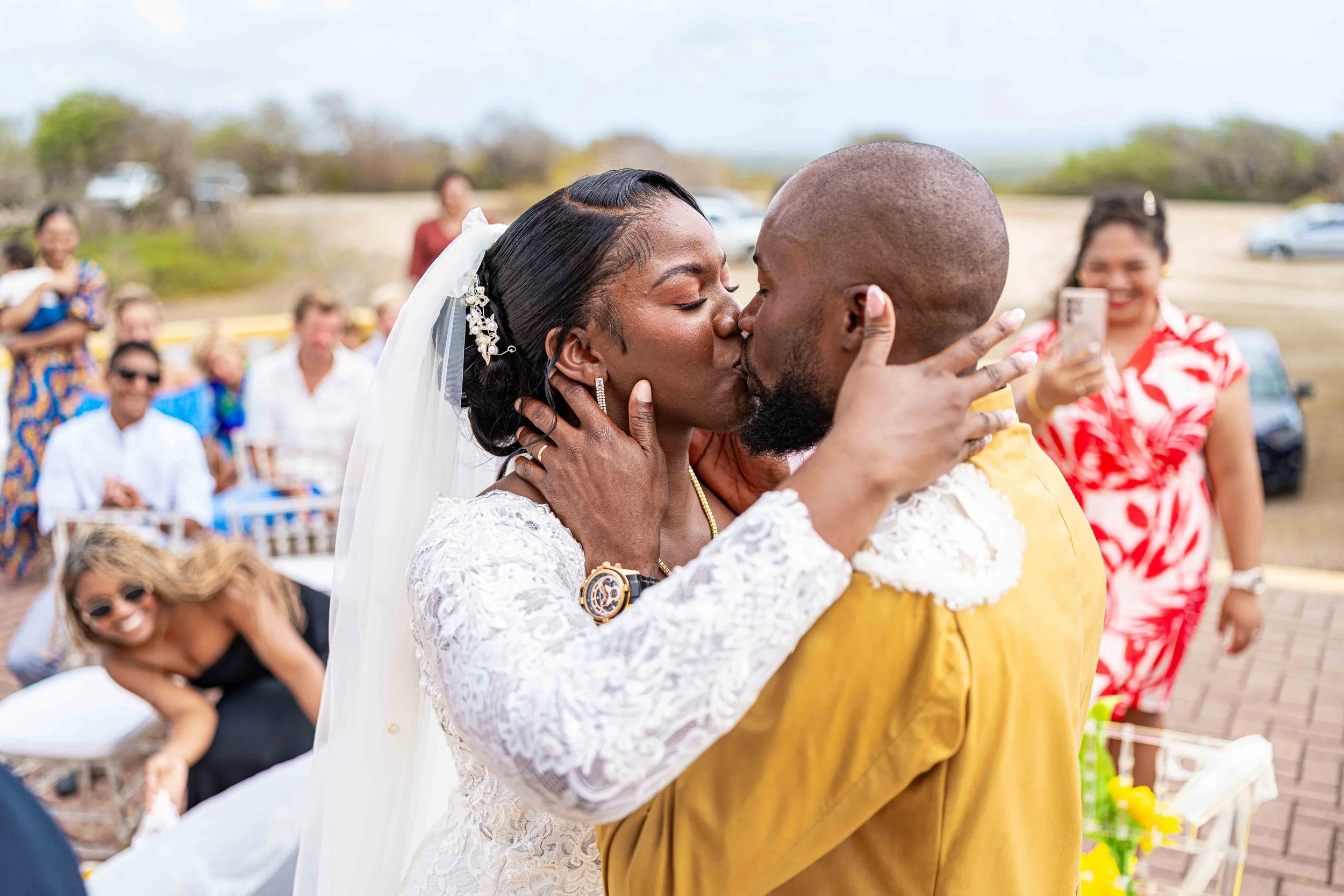 Een bruid en bruidegom kussen elkaar tijdens een buitenceremonie, omringd door vakantiegangers die de bruiloft vastleggen.