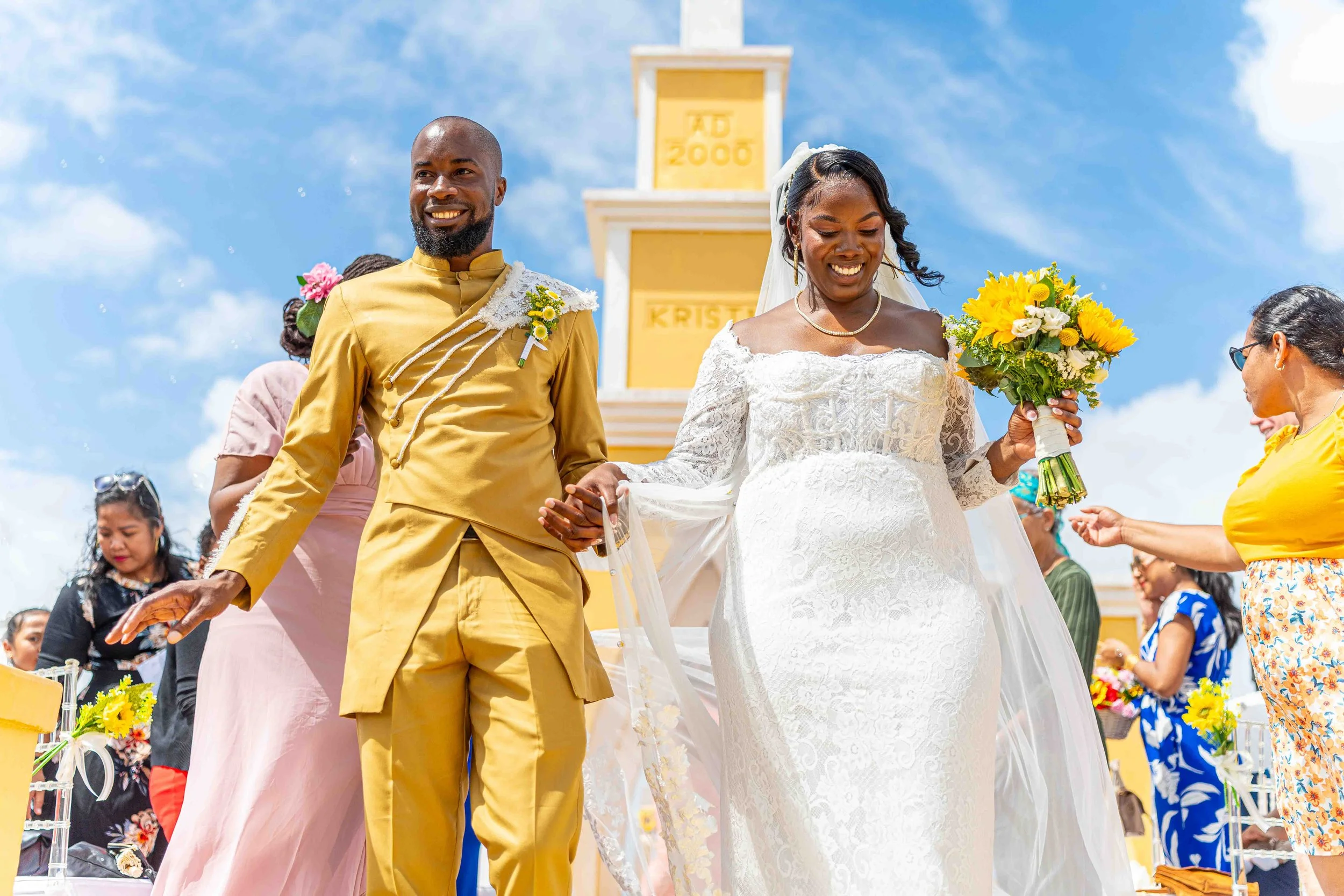 Een bruid en bruidegom lopen hand in hand tijdens hun huwelijksceremonie buiten, met een heldere blauwe lucht en witte wolken op de achtergrond.