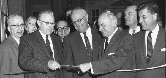 Black-and-white photograph of seven men in suits at a ribbon-cutting ceremony in 1960