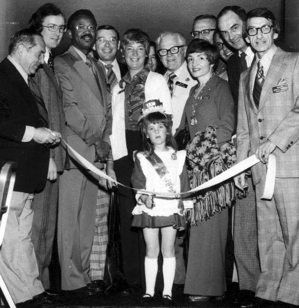 Black-and-white photograph of the 1975 WESTPEX ribbon-cutting ceremony with six-year-old Miss WESTPEX Tara Pope dressed in Irish costume, surrounded by WESTPEX volunteers and dignitaries
