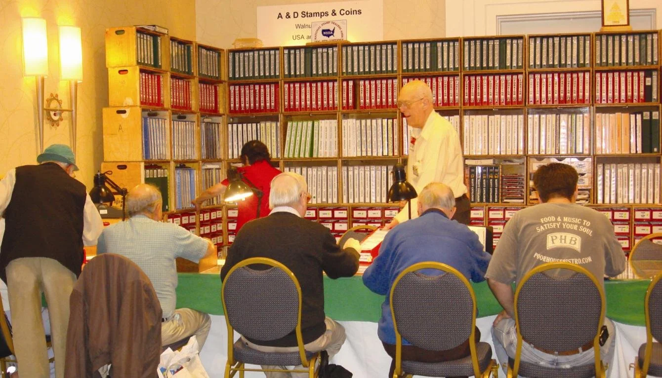 The A&D Stamps & Coins booth at WESTPEX with all ten chairs occupied and customers browsing the 'great wall' of stock books