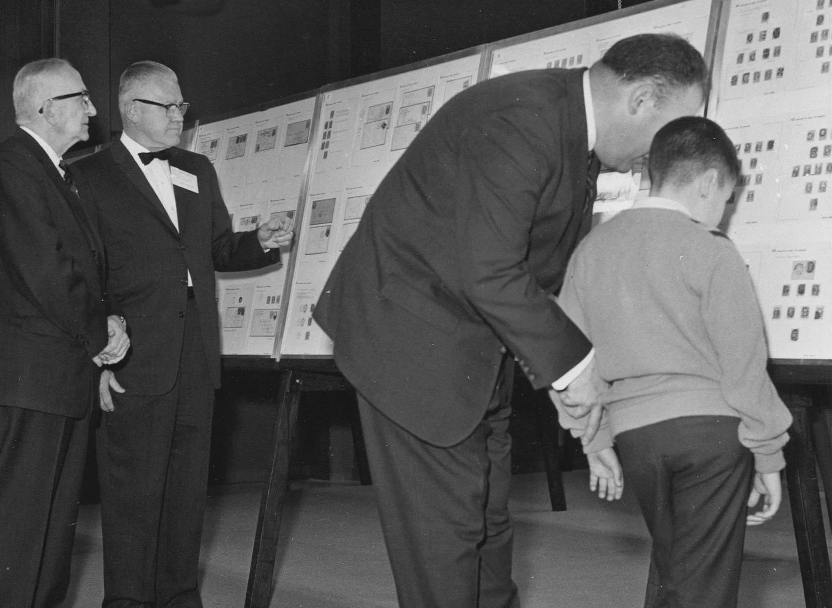 A black-and-white photograph from 1963 showing a father and son examining stamp exhibits, with Fred Thomas and Joseph Clary in the background