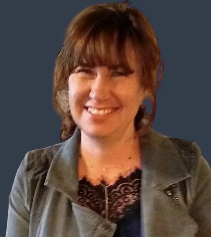 A woman with brown hair smiling, wearing a leather jacket and a necklace against a dark blue background.