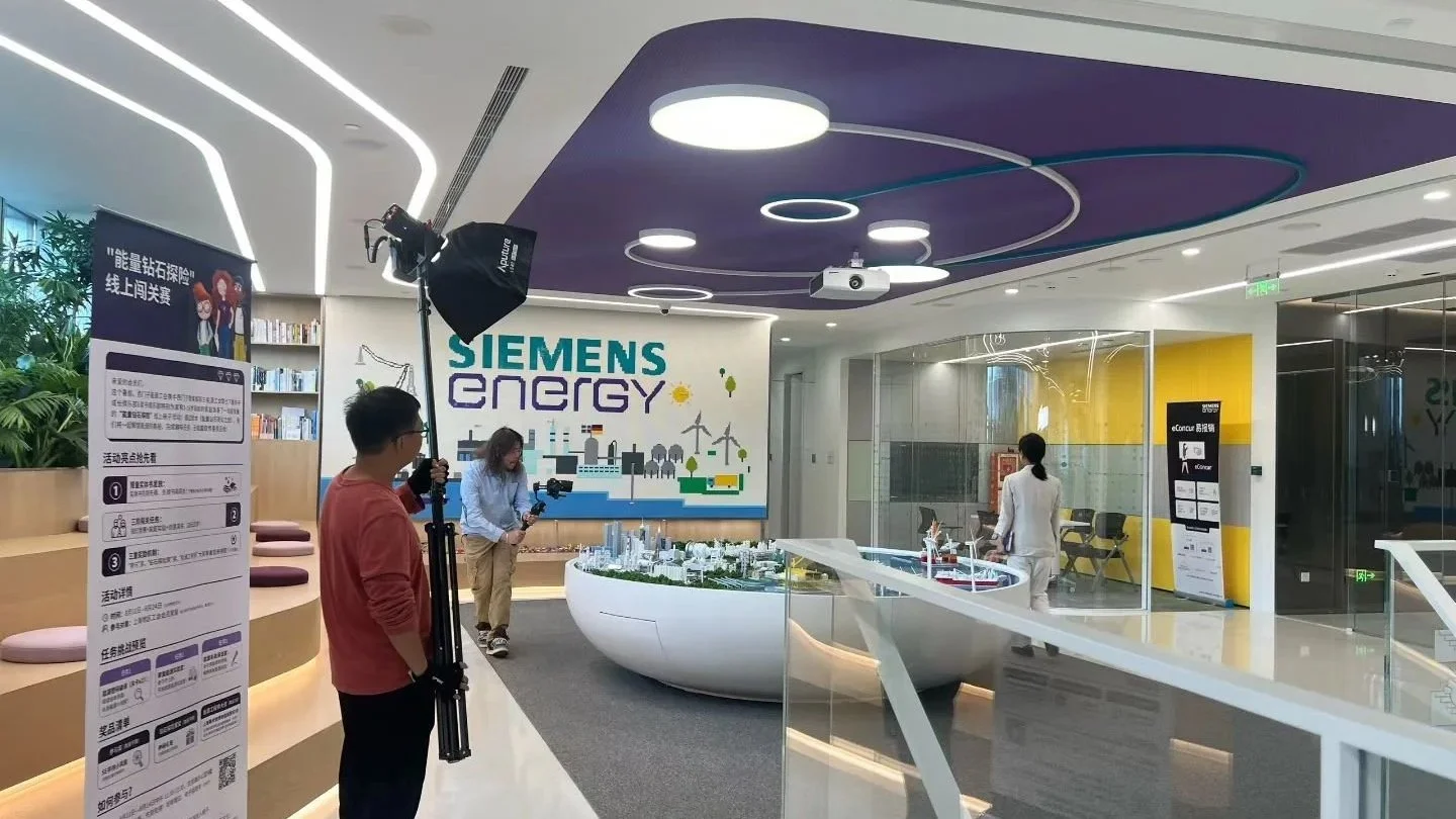 Inside a modern office building or corporate space with a Siemens Energy sign. There is a large model of a city or industrial area, a camera crew with a person holding a camera on a tripod, a woman in a white suit walking, and another woman setting up equipment. The area features sleek design elements, purple ceiling accents, and bright lighting.