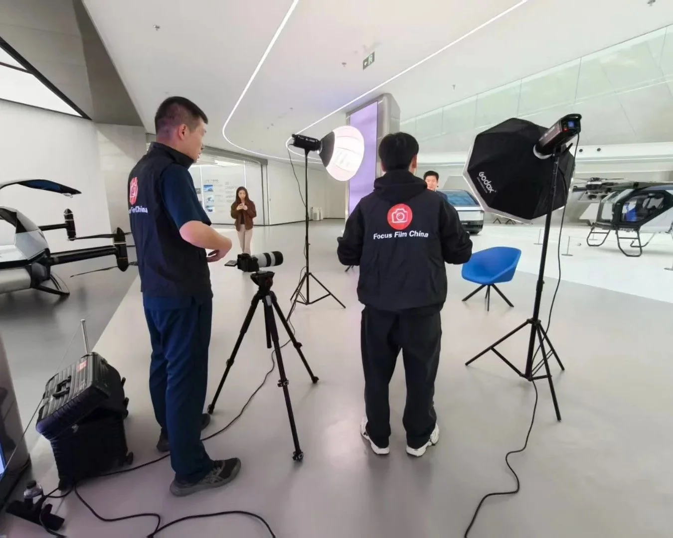 Two photographers setting up lighting equipment in a modern, spacious showroom with aircraft and helicopters in the background. One photographer is adjusting his camera, while the other stands nearby, both wearing jackets with 'Focus Film China' logo.