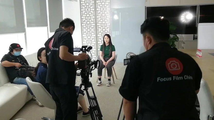 People filming a woman sitting against a wall in a room with large windows and modern decor.