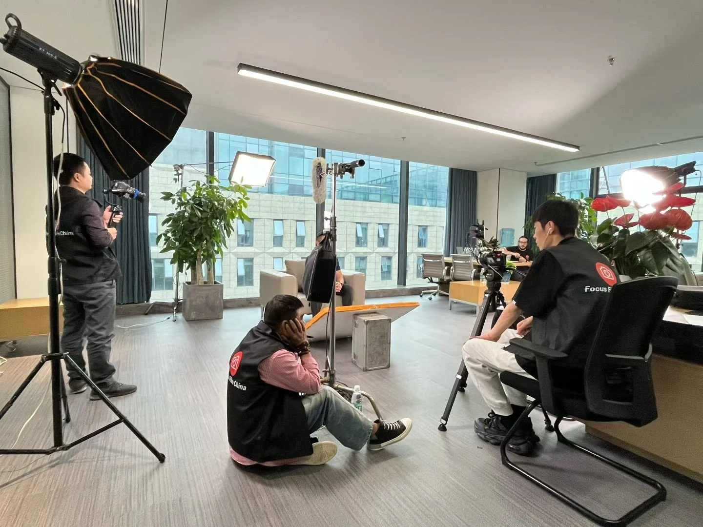Interview setup in an office with a person sitting on a couch being filmed by a camera crew, with lighting equipment and crew members preparing for the shoot.