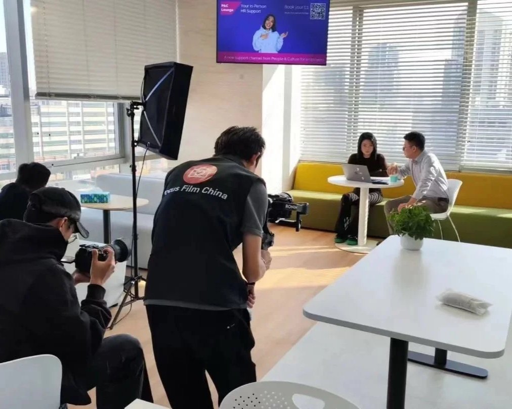 Two people are sitting at a small round table in a brightly lit room, working on a laptop. They are inside a modern office or lounge area with large windows and horizontal blinds. In the foreground, three photographers are capturing the scene, with one holding a camera and the others positioned near equipment. There is a large screen on the wall displaying a promotional message.
