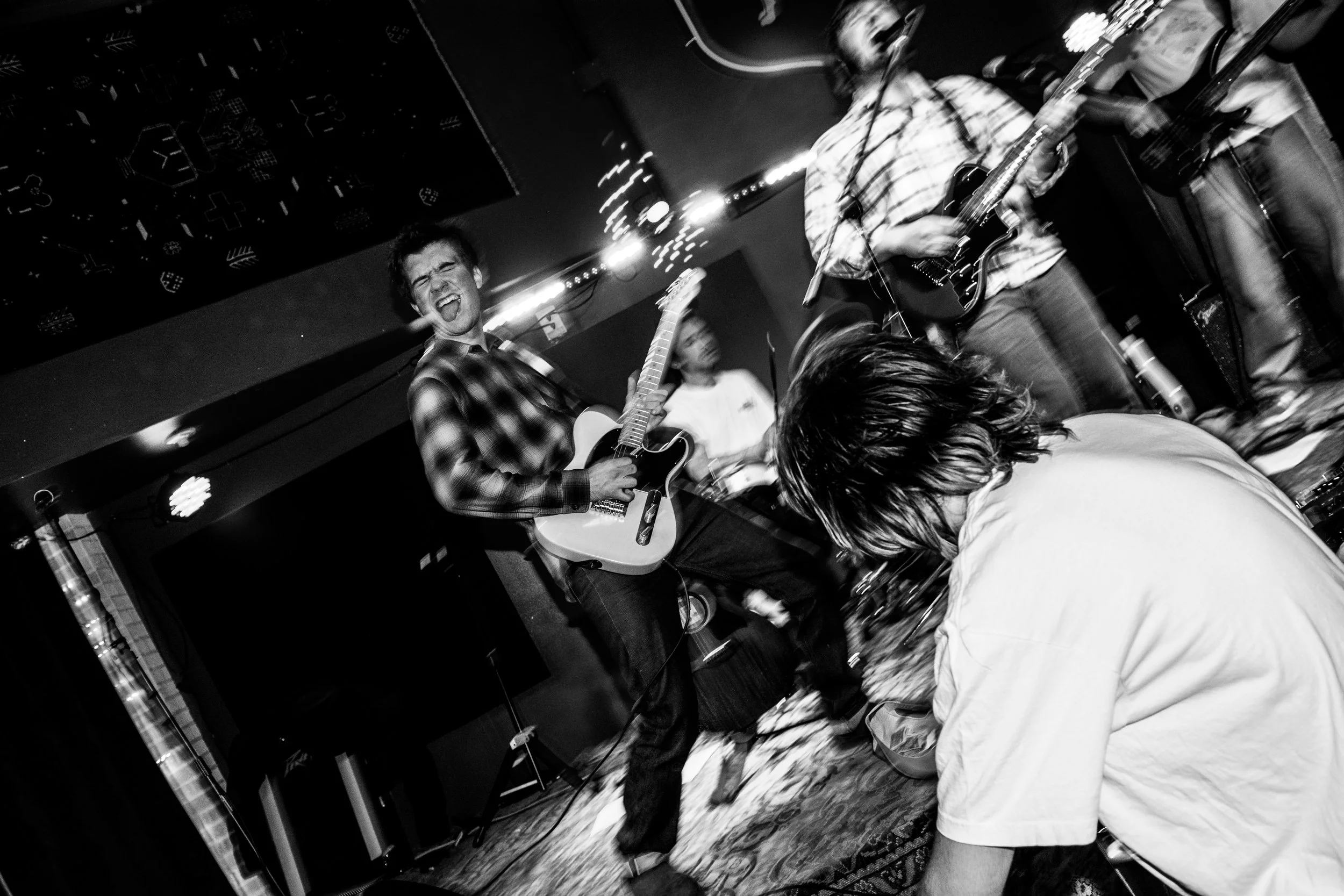 A band performing on stage with one member passionately singing while playing an electric guitar, and another playing an acoustic guitar, with a person in front crouching down or kneeling.