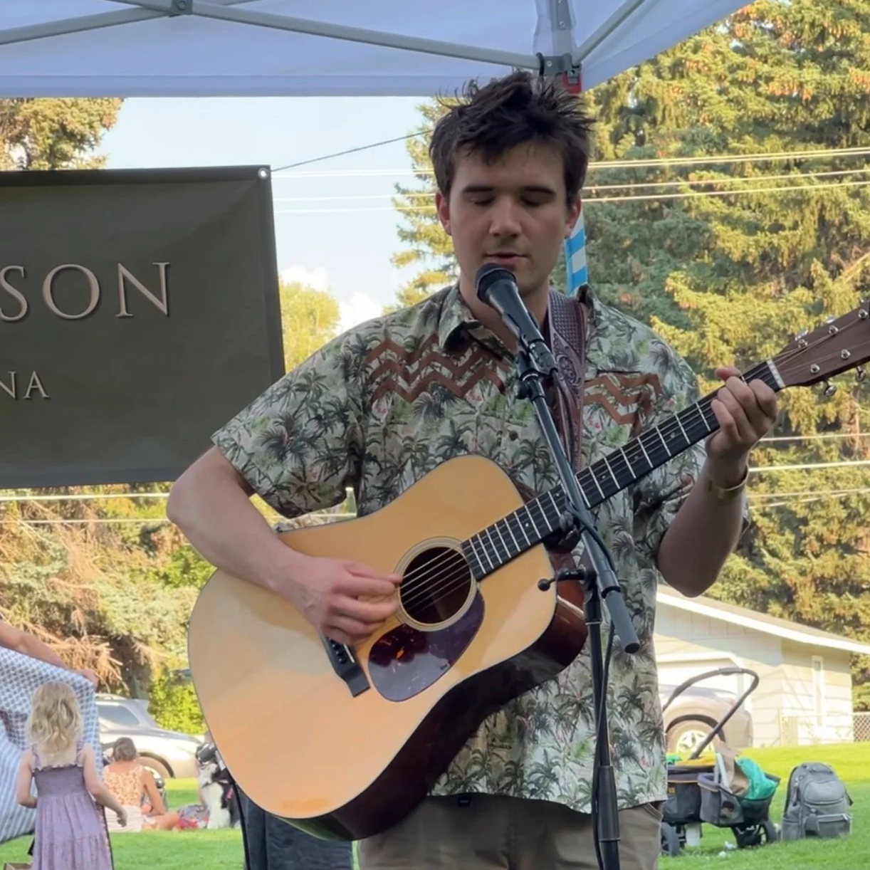 Young man playing an acoustic guitar and singing into a microphone at an outdoor event with trees and a house in the background.