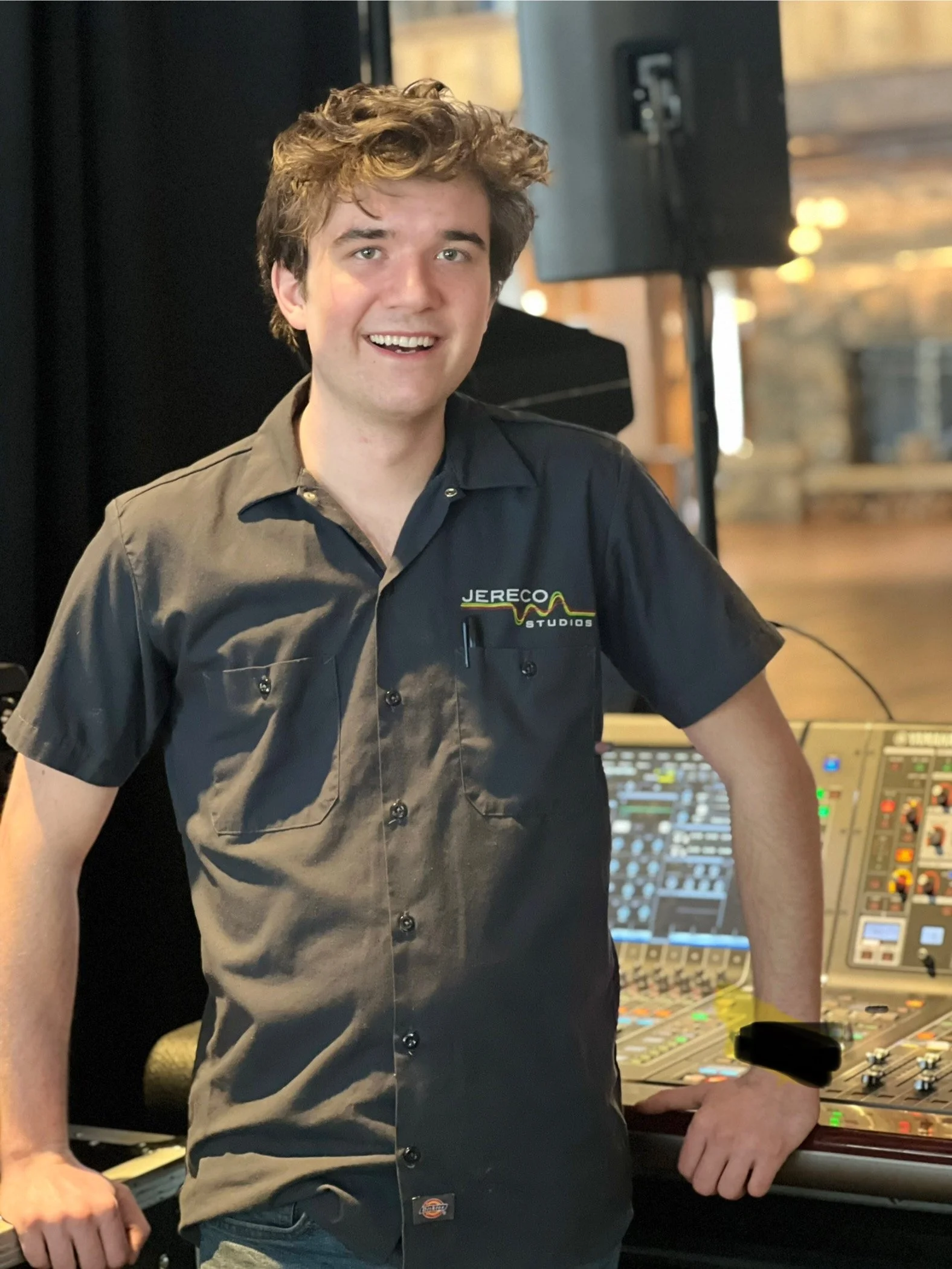 Young man smiling, wearing a black shirt with 'JERECO STUDIOS' logo, standing next to sound equipment in a recording or studio setting.