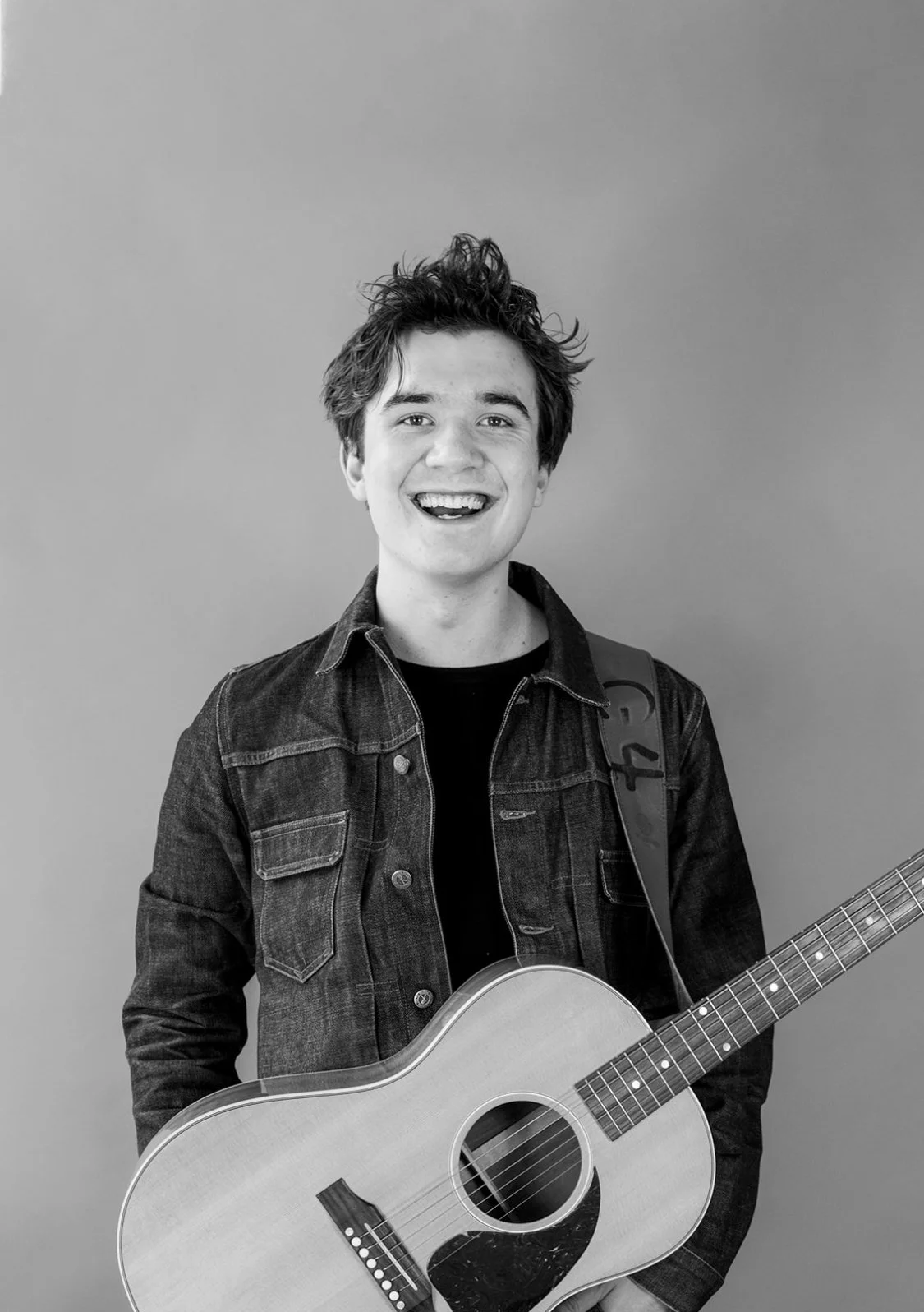 A young man with messy hair smiling and holding an acoustic guitar, wearing a denim jacket, standing against a plain background.