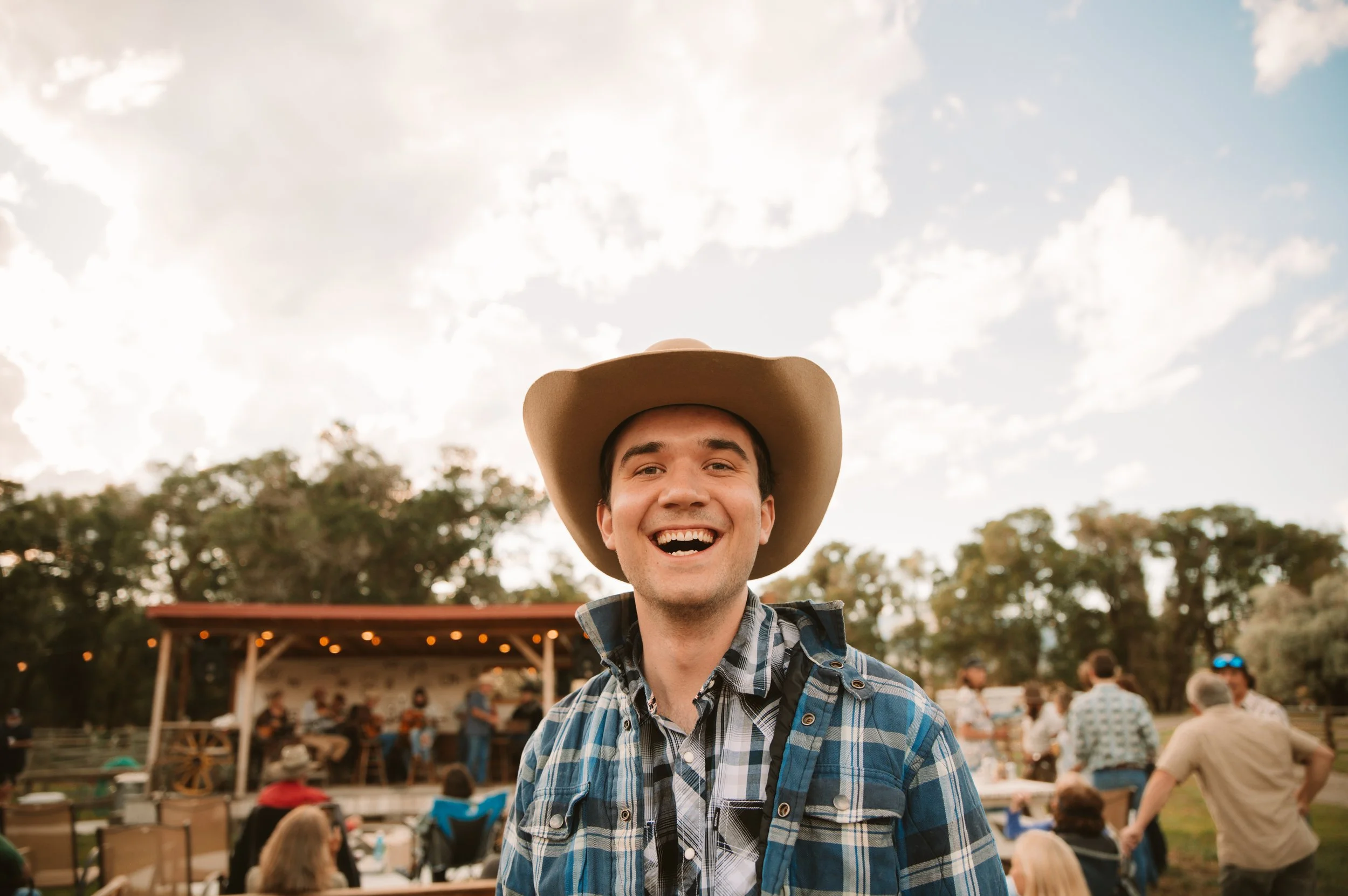 A man wearing a cowboy hat and plaid shirt smiling at an outdoor event with people and a stage in the background under partly cloudy sky.