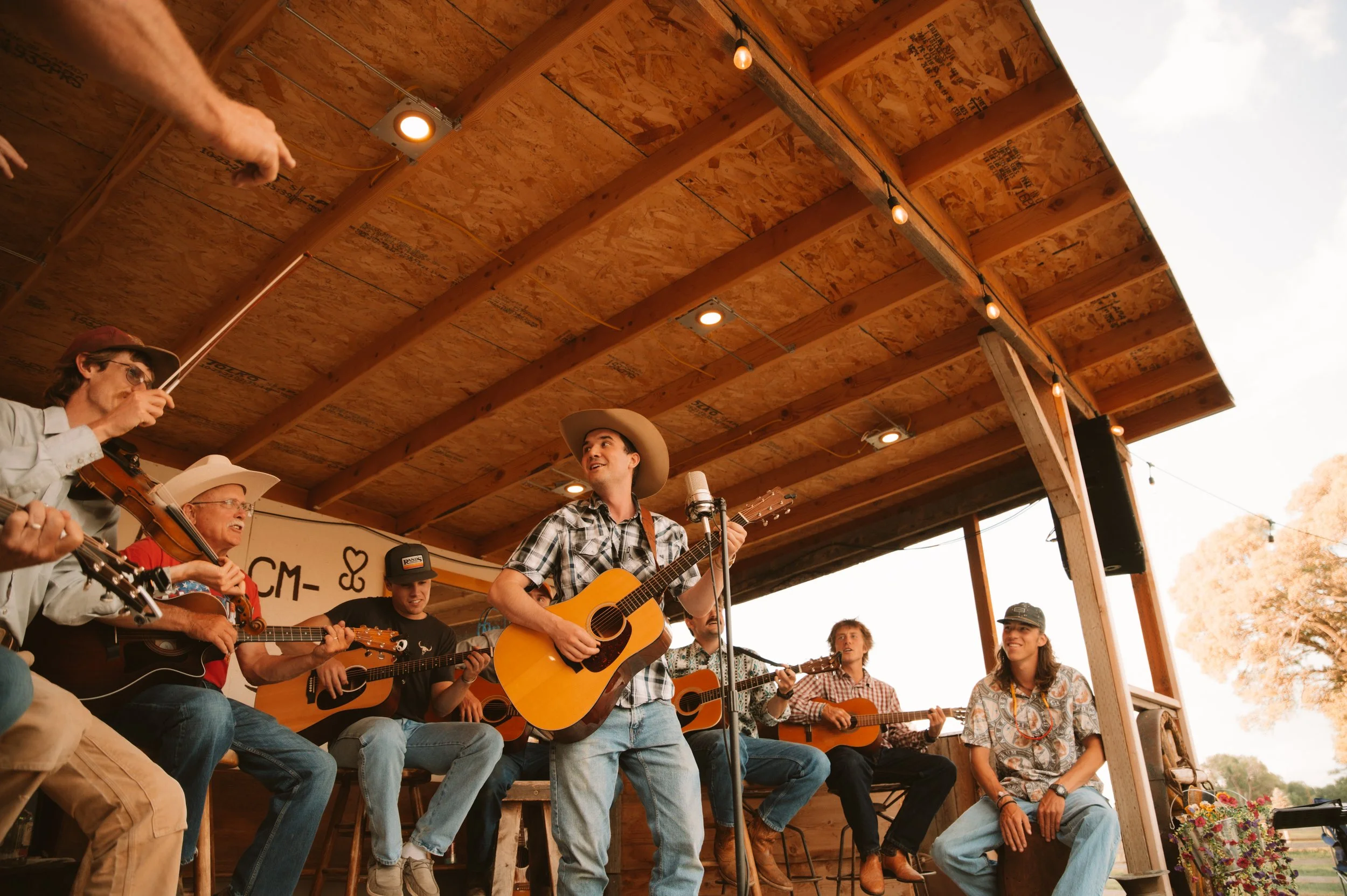 Group of men and women playing guitars and singing on stage under a wooden roof at an outdoor event, with some wearing cowboy hats and casual clothing.