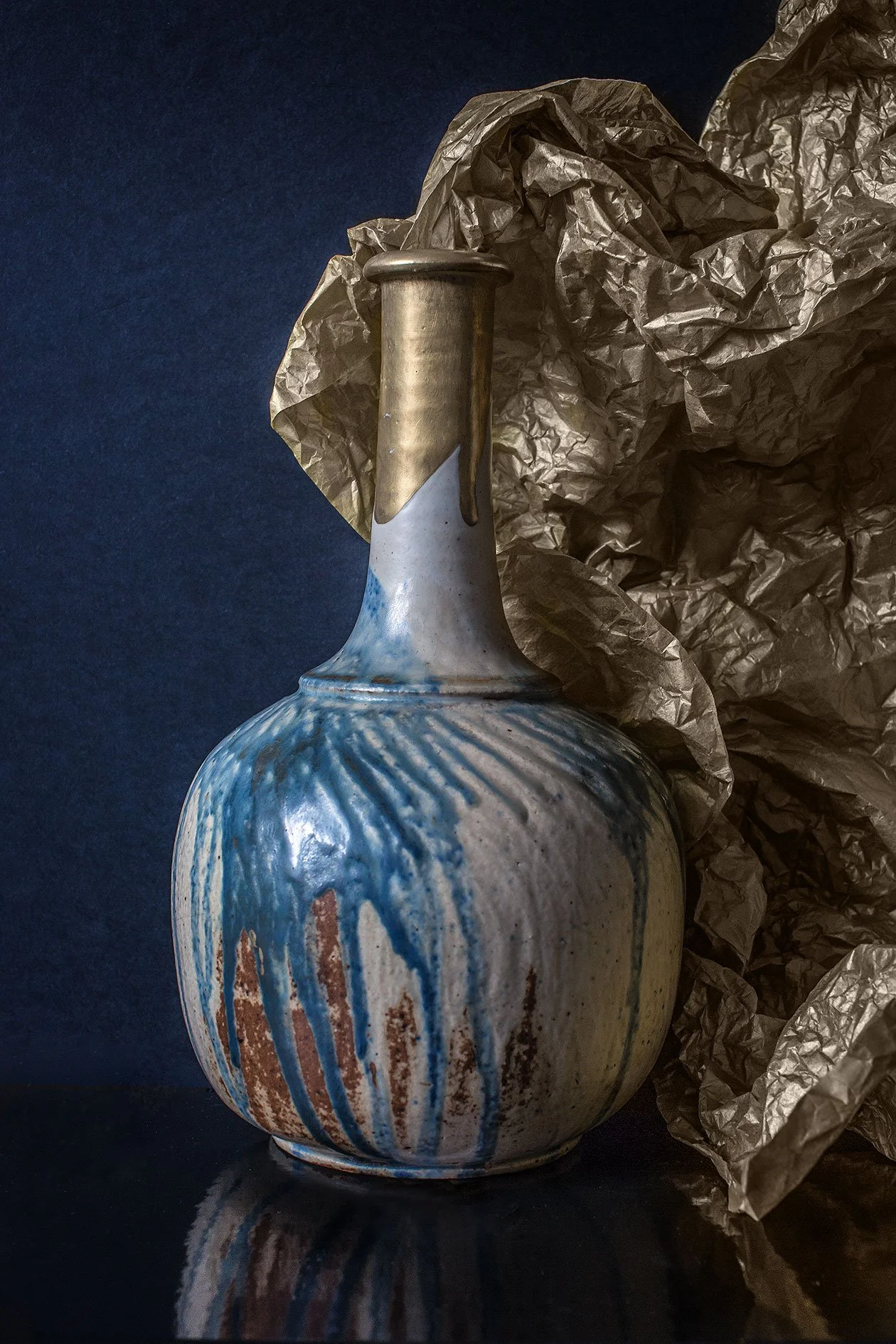 A ceramic vase with blue and brown vertical streaks, placed on a dark surface, with crumpled brown paper in the background.