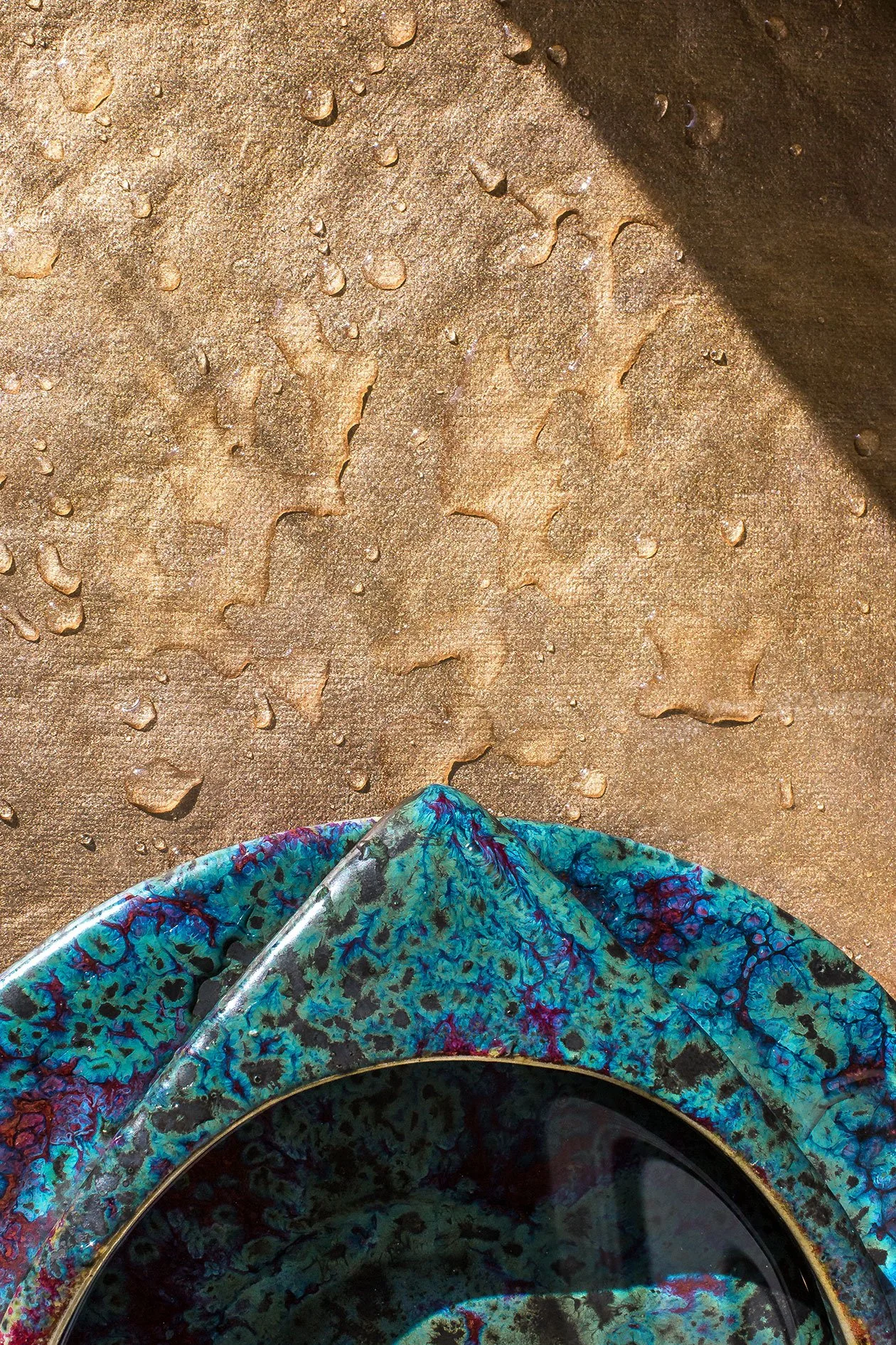 Close-up of a decorative, textured earrings with a swirling blue and purple pattern, resting on a fabric with water droplets on a textured, sandy or bronze-colored surface.