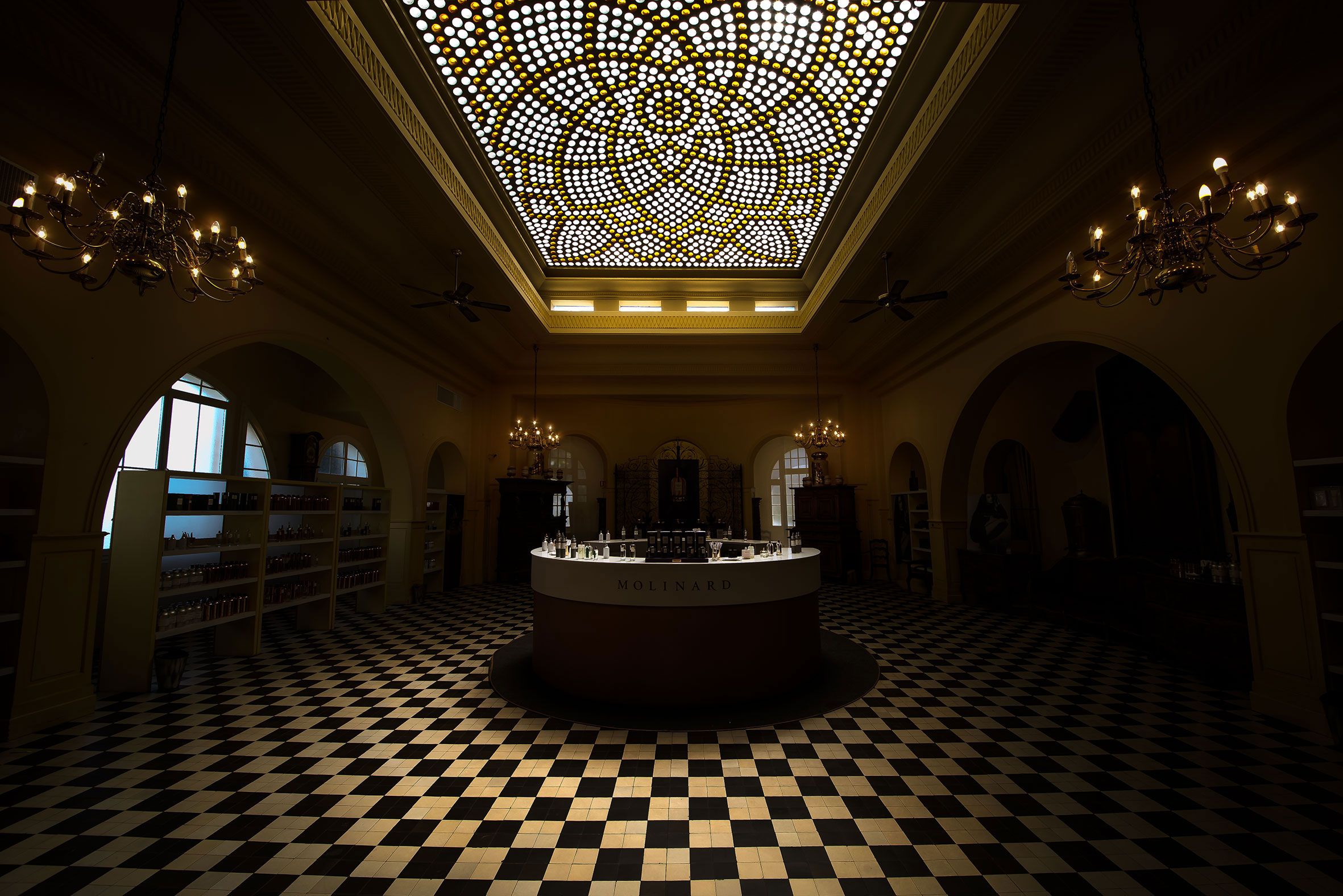 Interior of an elegant room with a patterned black and white checkered floor, chandeliers, and a large decorative ceiling light panel with yellow and white lighting. A circular display table with bottles is in the center, labeled 'MOLINARD', and arch