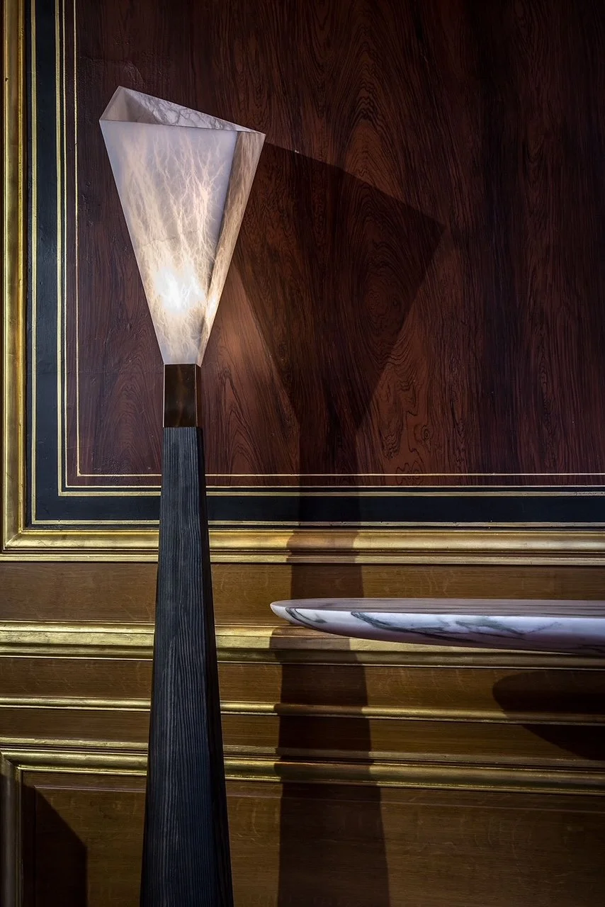 A tall, modern lamp with a geometric, cone-shaped shade and a black base, placed on a wooden surface, with an ornate wooden wall and a marble-lined shelf in the background.