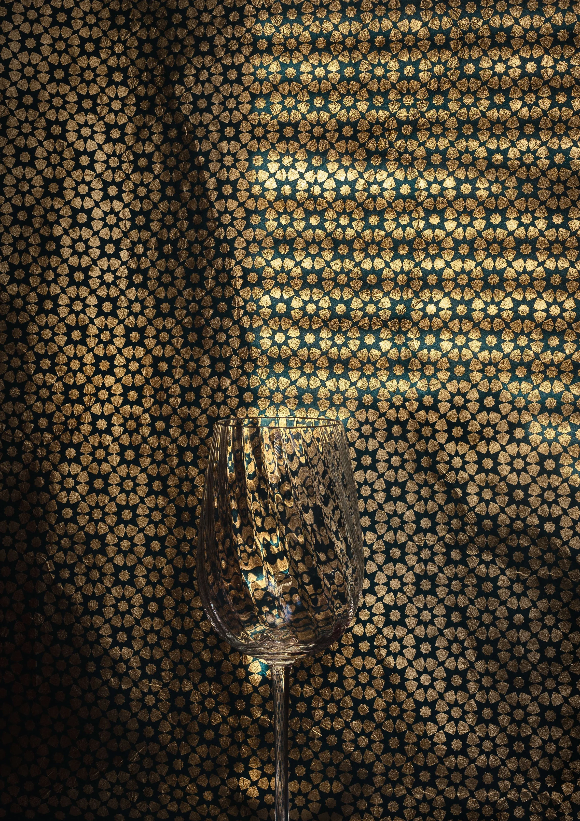 A clear glass goblet with a wide bowl, slender stem, and a reflective surface, placed on a patterned surface with a geometric star design in gold on a black background.