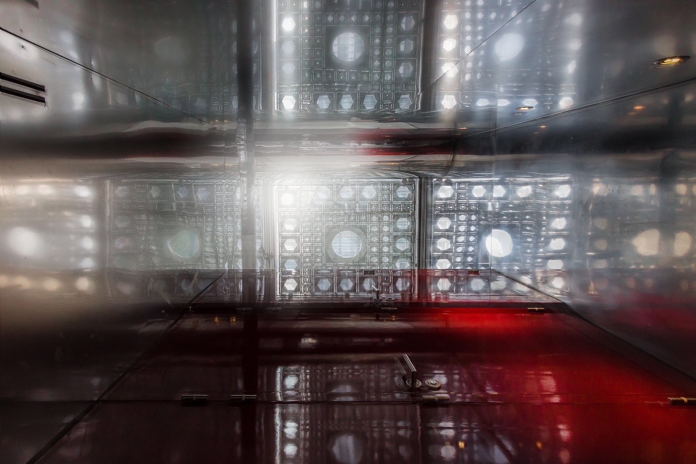 Looking up inside a metallic elevator shaft with light at the top and reflecting surfaces.