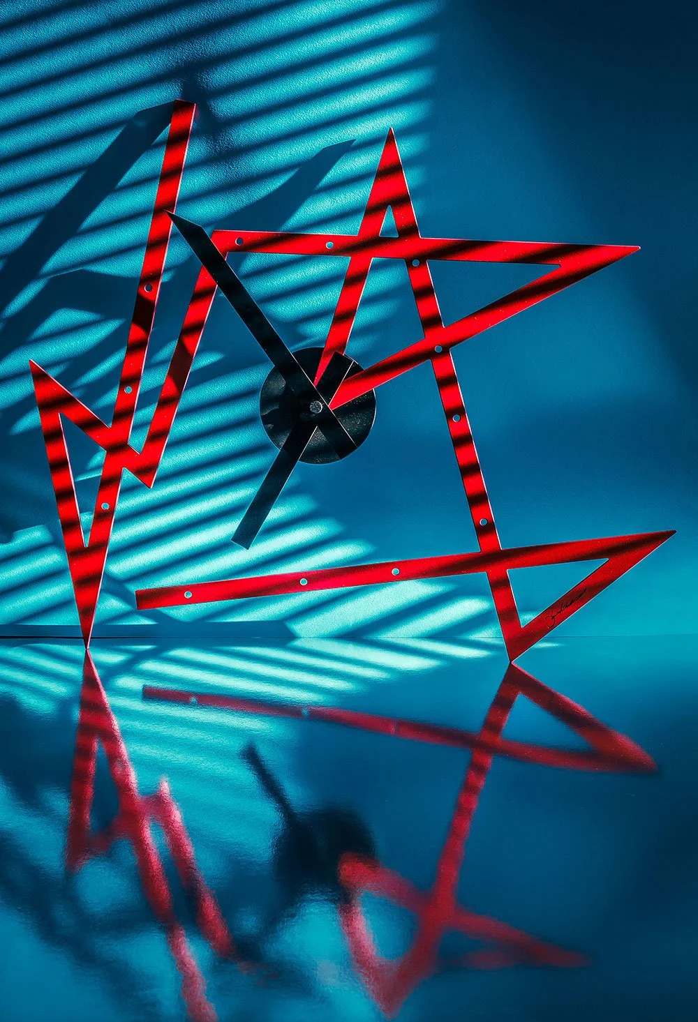 Modern wall clock with red geometric hands and markers, casting shadows on a textured blue wall with horizontal light stripes.