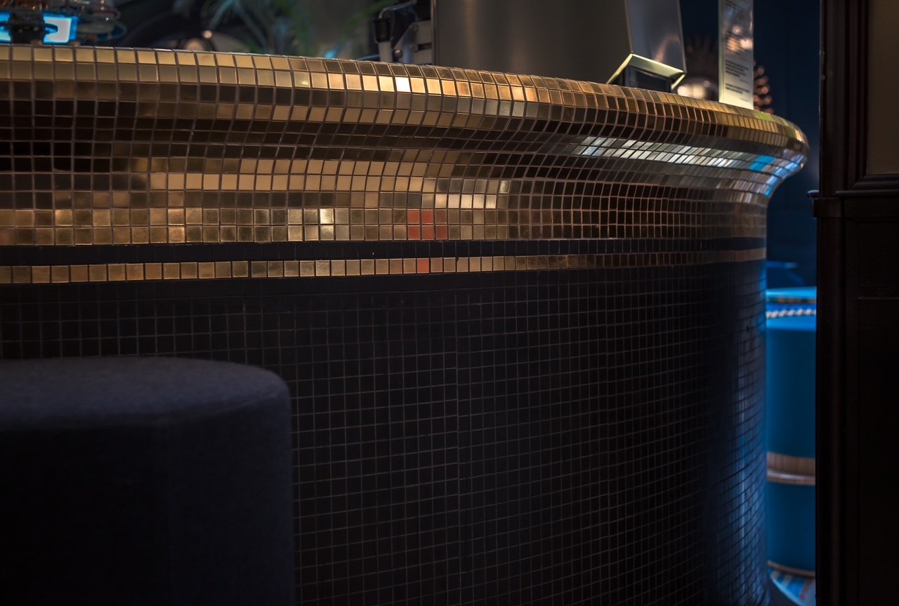 A close-up view of a curved bar counter with a mosaic tile pattern, and a dark seating area in the foreground.
