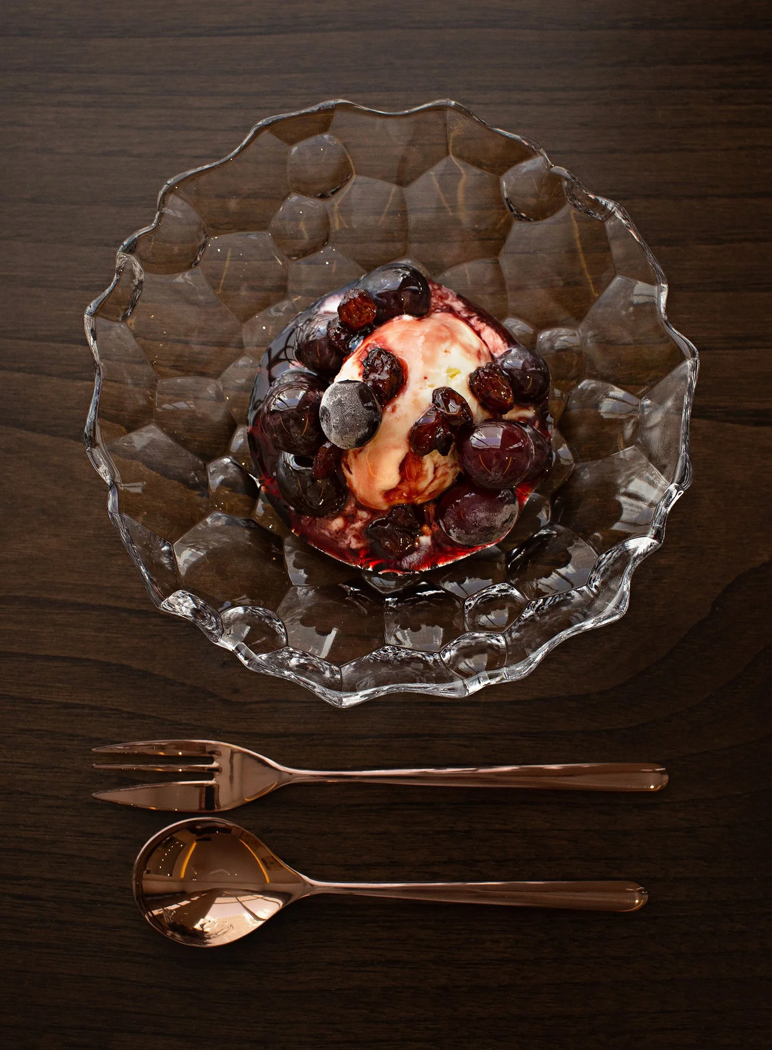 A glass bowl with a wavy edge containing a dessert topped with blueberries and a scoop of ice cream, placed on a wooden table with a rose gold spoon and fork nearby.