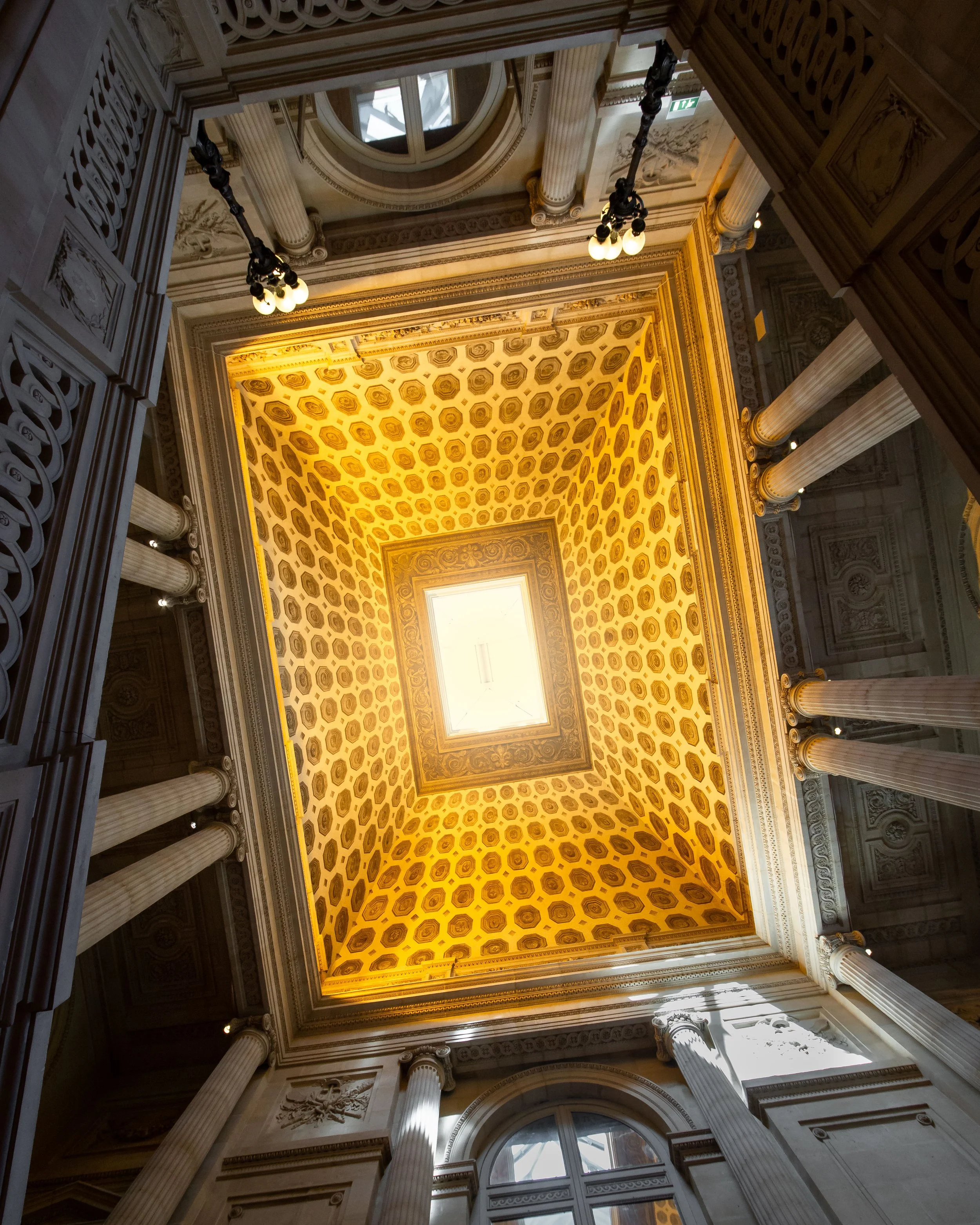 View upward inside a grand historic building with ornate architectural details, tall columns, a decorated ceiling with patterns, and a skylight.