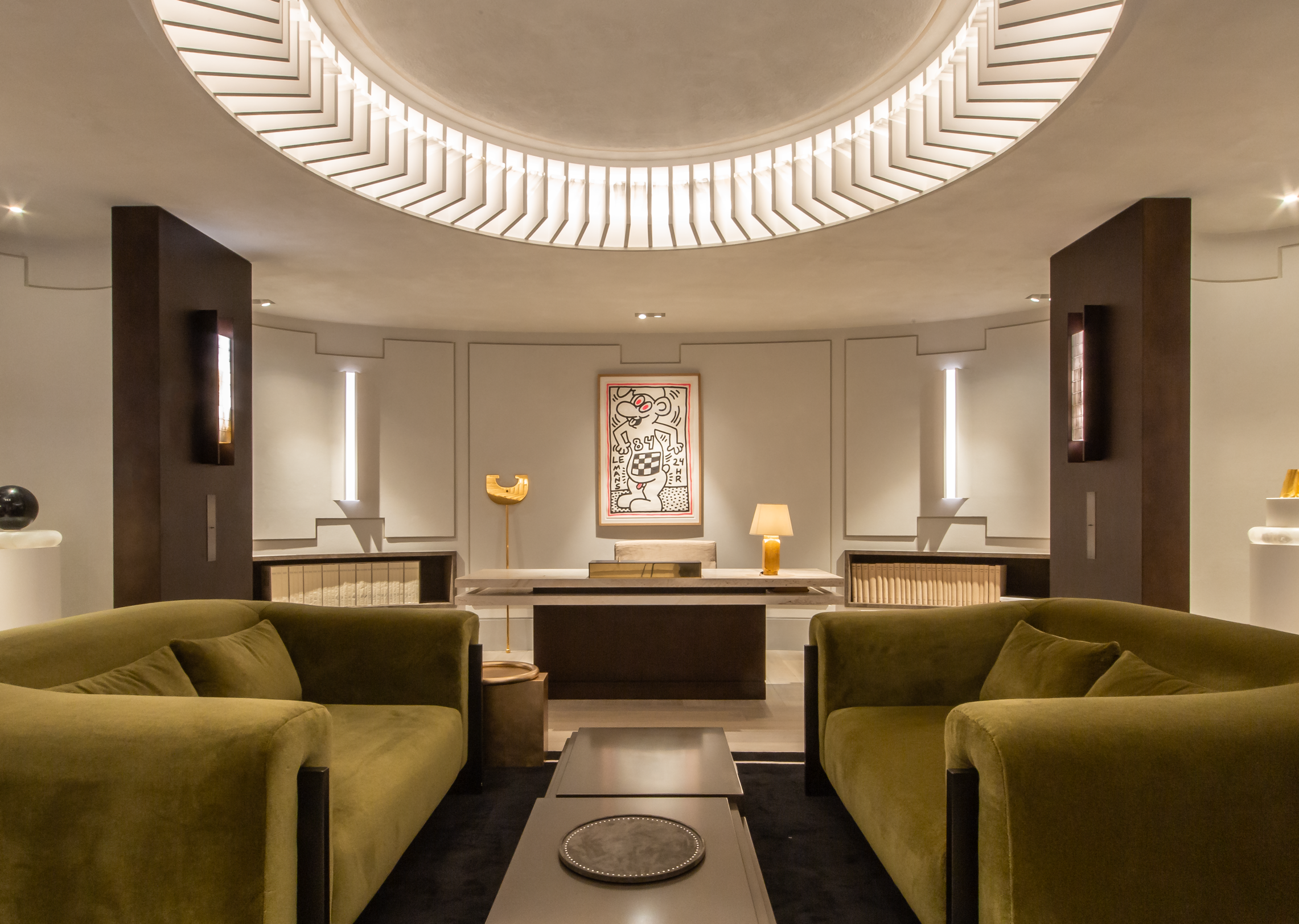 A spacious living room with two green sofas facing each other, a black coffee table in the center, and a large circular opening in the ceiling with modern lighting. There is artwork on the wall and some lamps providing ambient light.