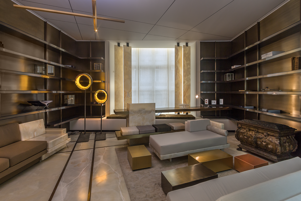 Modern hotel lobby with beige and white sofas, gold and bronze coffee tables, dark wooden wall shelves, marble flooring, and warm lighting.