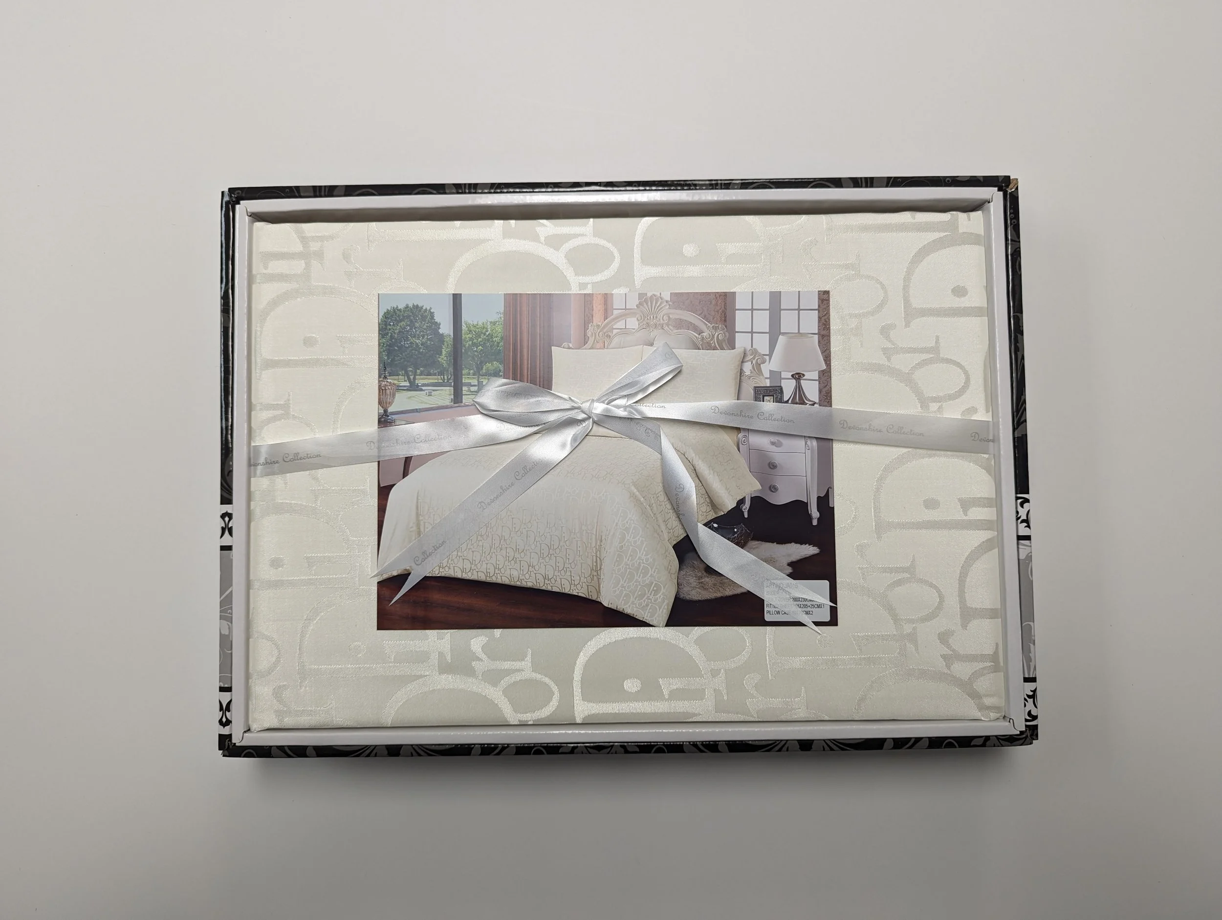 A boxed bedding set with a white satin ribbon, featuring a photo of a bedroom with a white bed, nightstand, lamp, and large window with green trees outside.
