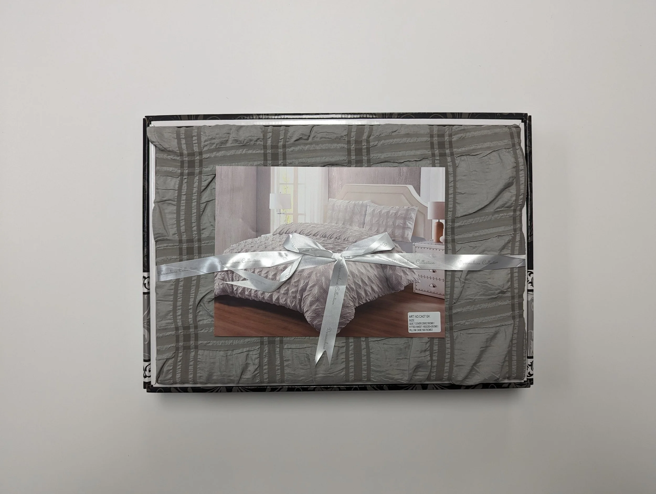 A boxed bedding set with a picture of a bed on the front. The box is wrapped with a silver ribbon and placed on a white surface.