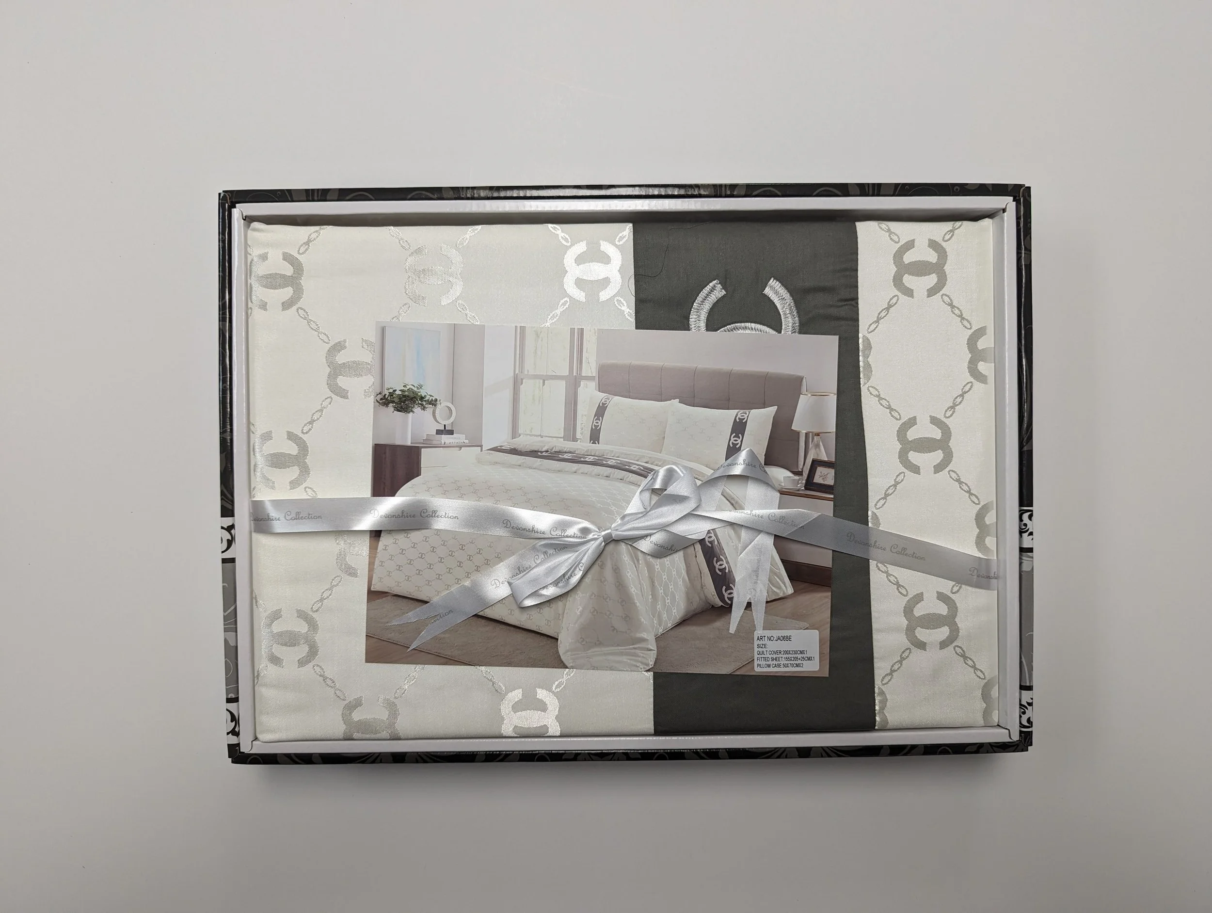 Packaging of a bedroom collection with a photo of a neatly made bed, white and grey bedding, a headboard, nightstand, and lamp, wrapped with a silver ribbon with some product information visible.