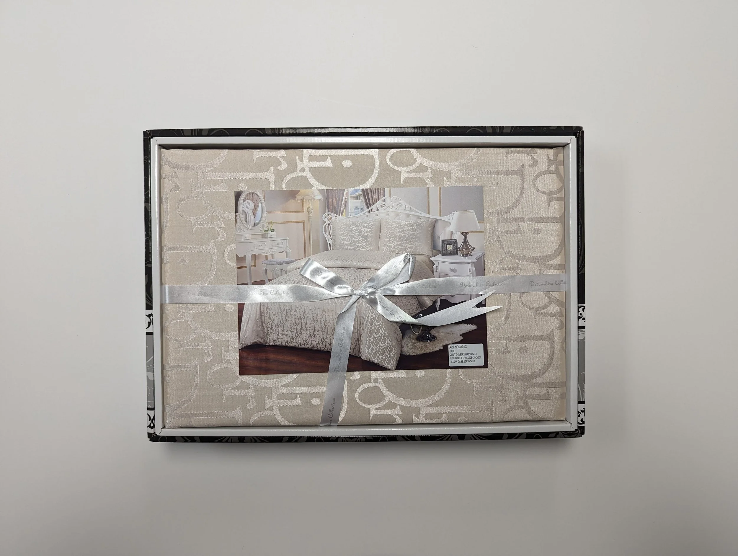 Box containing a white themed bedding set with a picture of a bedroom in the center, tied with a silver ribbon.