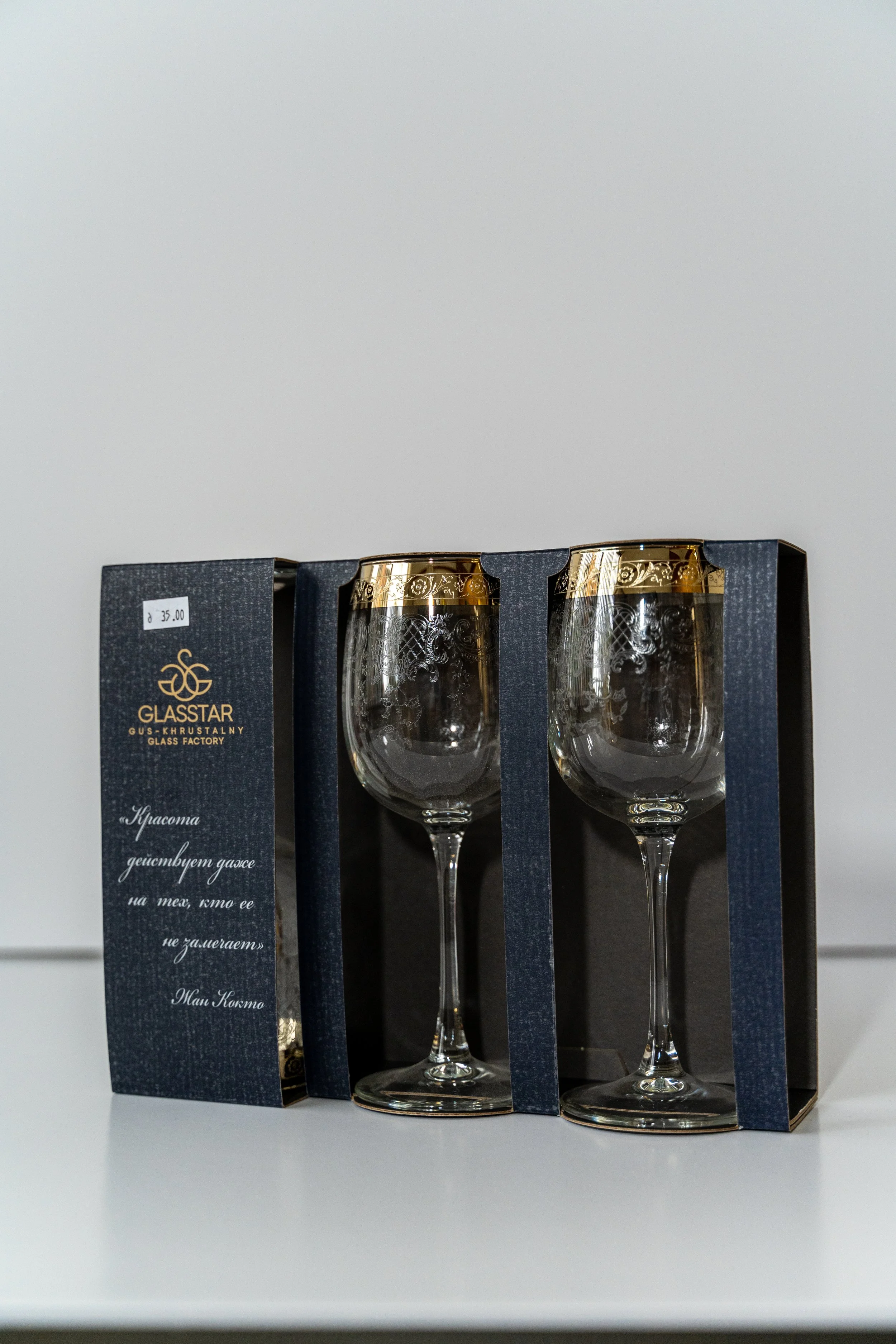 A set of two clear glasses with gold rims, packaged in a dark blue box from Glasttar, a glass factory, placed on a white surface.