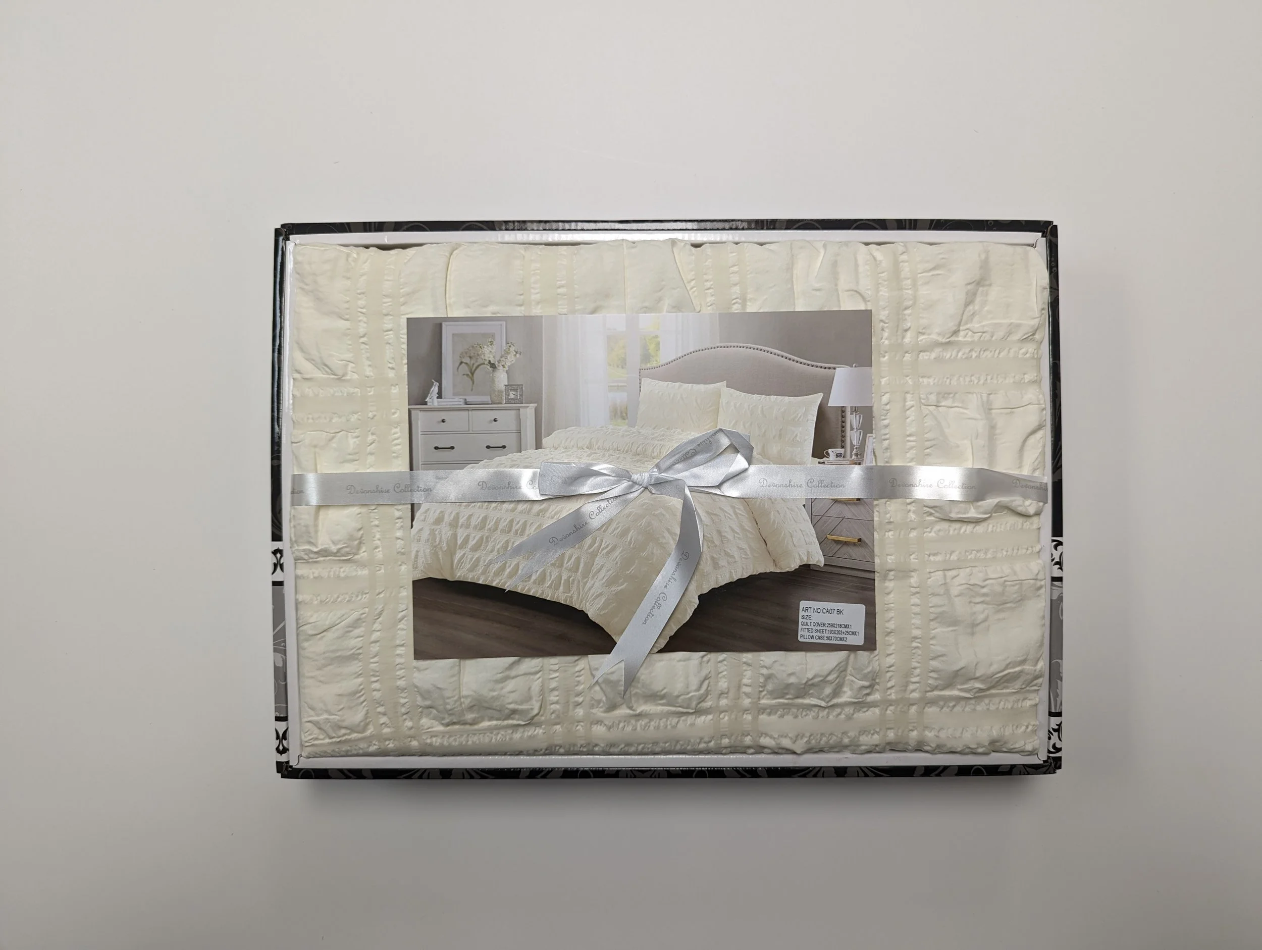 Box containing a cream-colored beddings set with a picture of a bedroom on the front, tied with a silver ribbon.