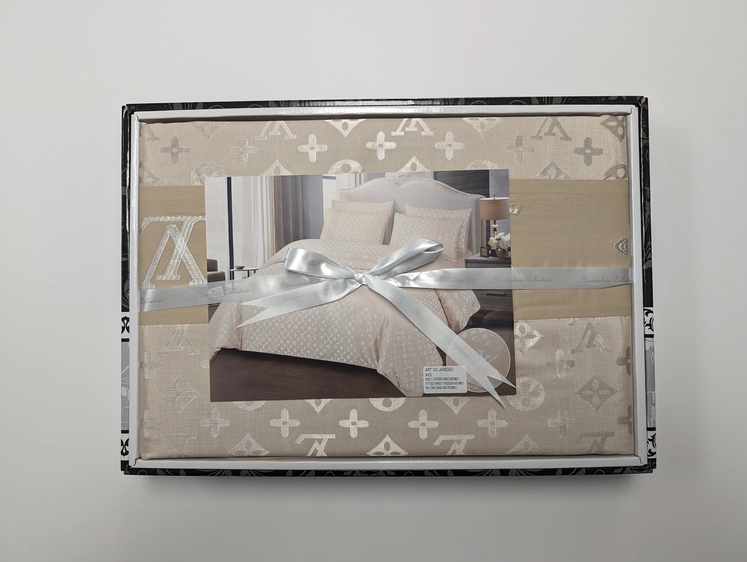The image shows a packaged bedding set with a picture of a neatly made bed with beige and white patterned bedding and pillows, wrapped with a silver ribbon.