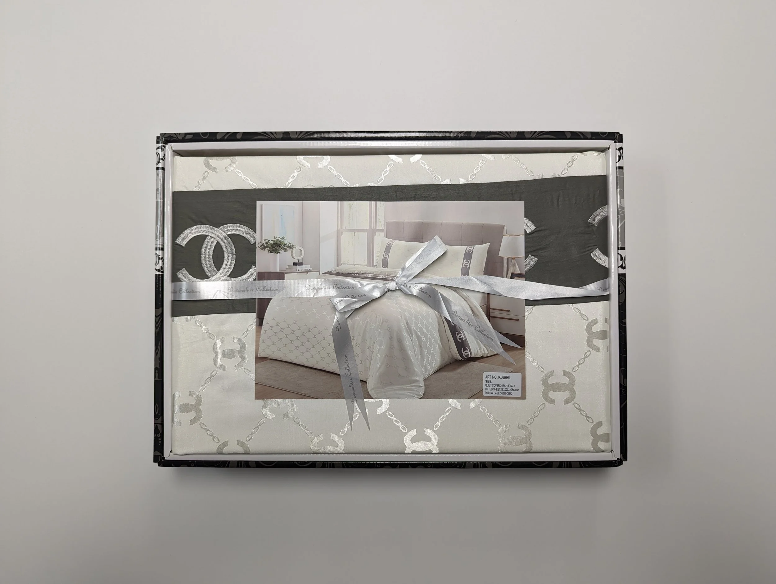 Box containing a bed set with white and gray bedding, tied with a gray ribbon, featuring a bedroom scene with a gray headboard and cream pillows.