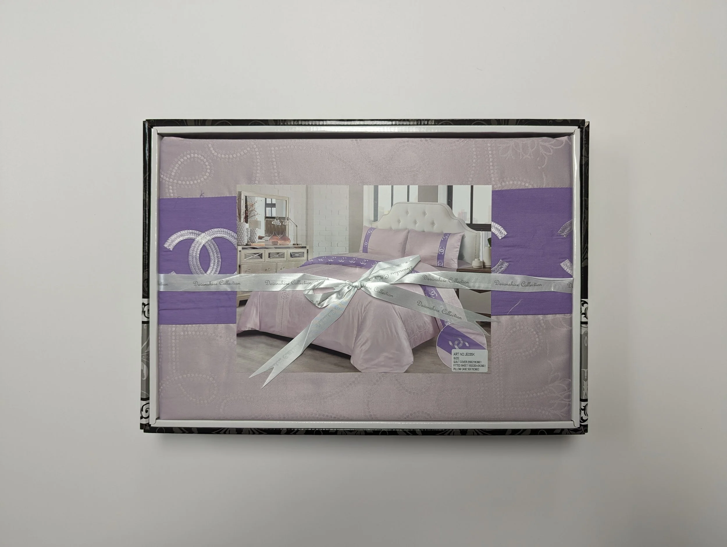 A boxed set of bedding with a bedroom image on the front, featuring a bed with a tufted white headboard, lavender and white bedding, and a dresser in a bright, modern room. The box is wrapped with a silver ribbon.